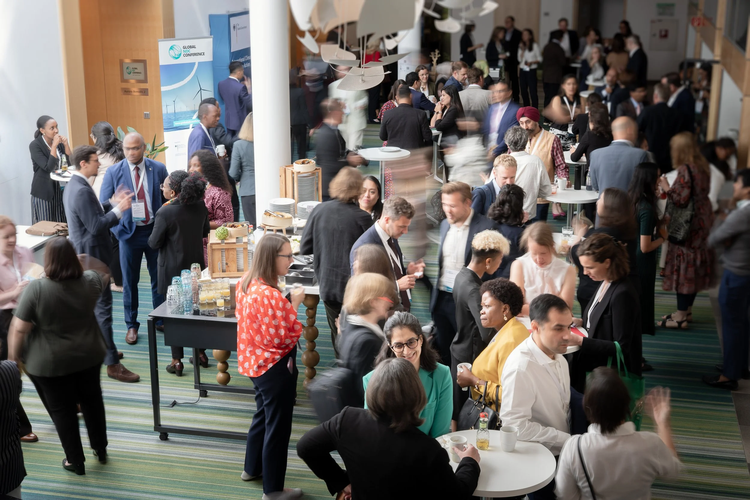 Ein großer, geschäftiger Konferenzraum mit vielen Menschen, die sich unterhalten, Getränke trinken und an Tischen essen. Im Hintergrund sind Banner und Informationsstände für eine globale Konferenz sichtbar.