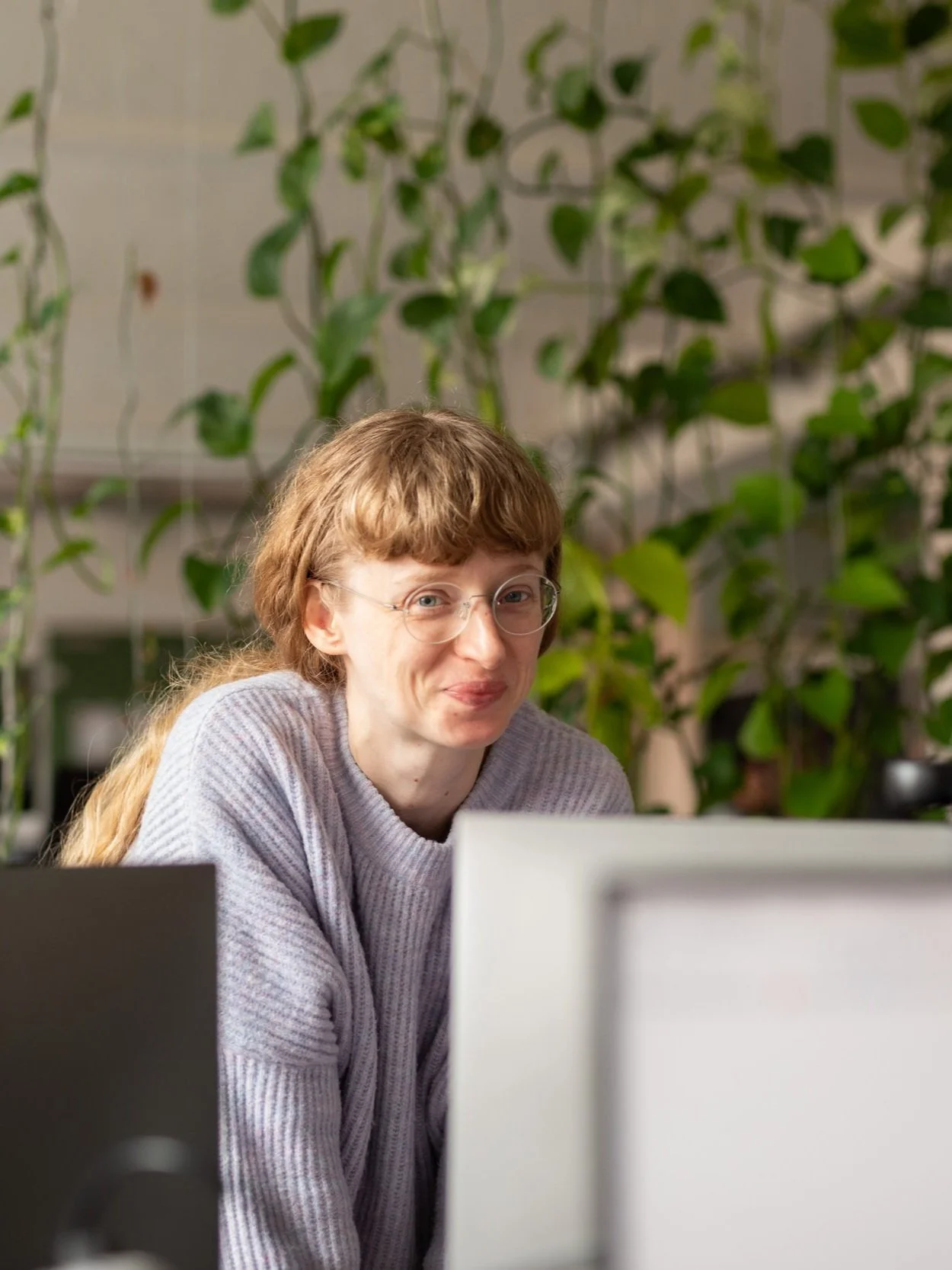 Eine Frau mit Brille und langen, lockigen Haaren, die an einem Computer arbeitet, im Hintergrund grüne Pflanzen.