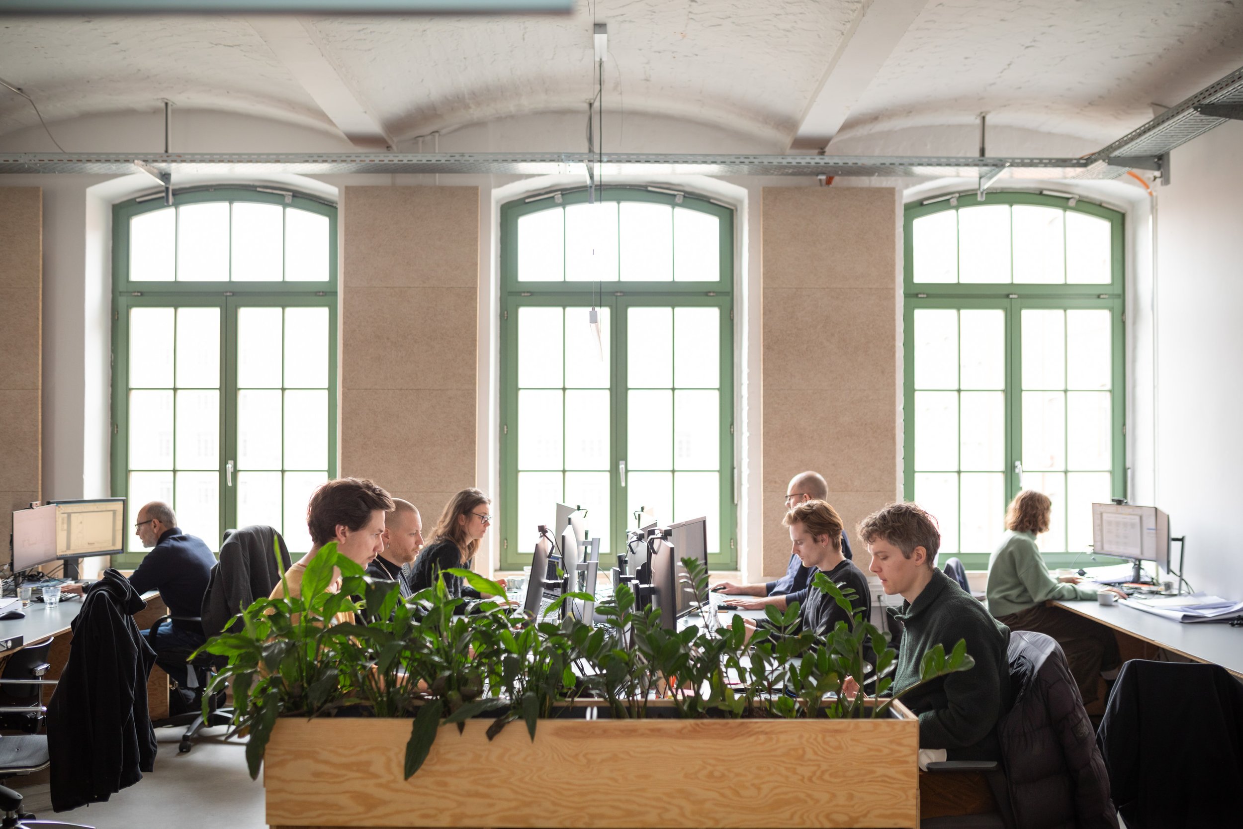Büroarbeitsplatz mit mehreren Personen, die an Computern arbeiten, große Fenster im Hintergrund, grüne Pflanzen im Vordergrund.