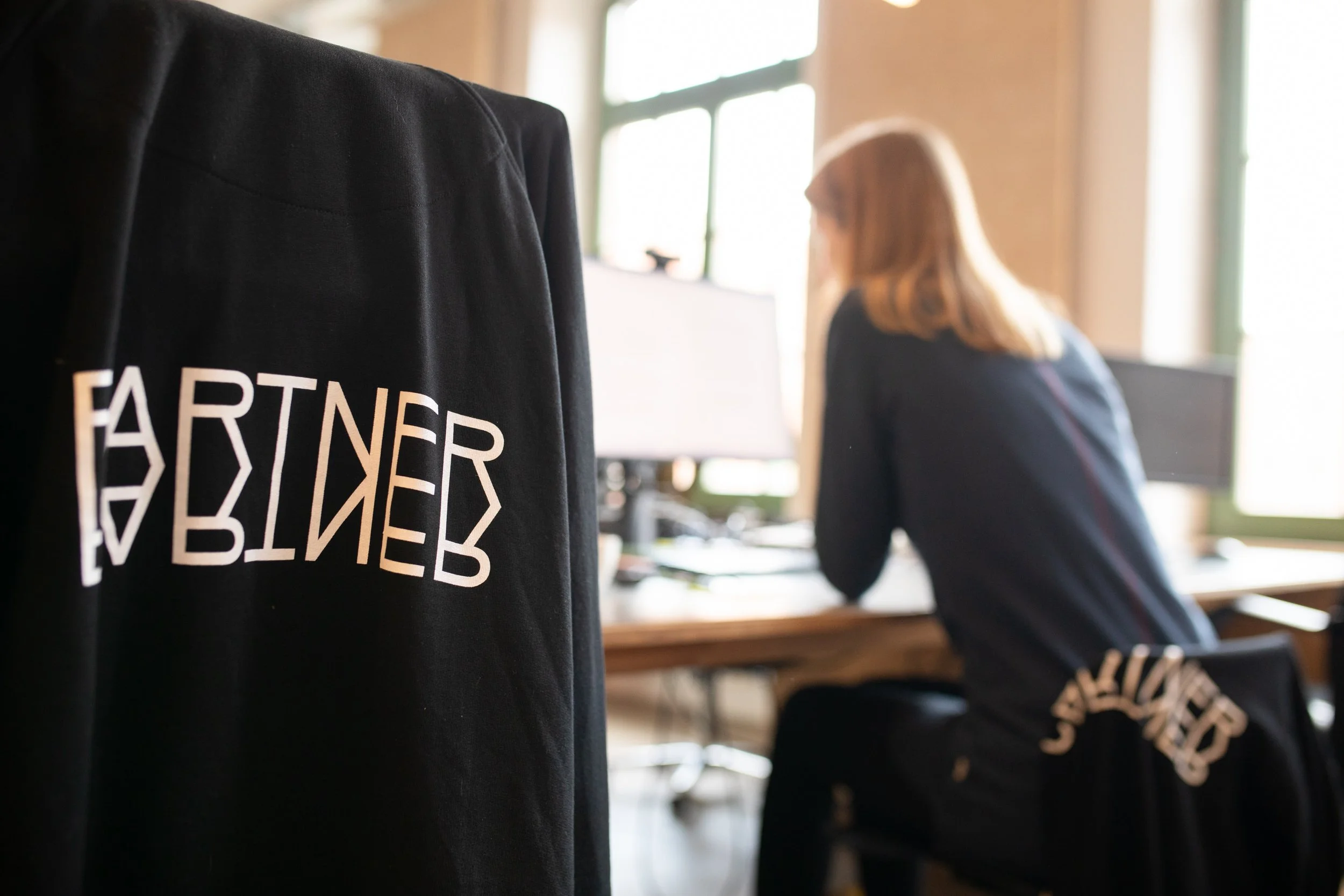 Nahaufnahme eines schwarzen Gedenk- oder T-Shirt mit weißen Schriftzug, im Hintergrund eine blondhaarige Frau. Das Bild ist unscharf, der Fokus liegt auf dem Text auf dem Kleidungsstück.