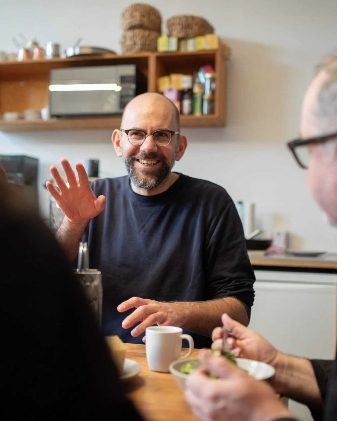 Ein Mann mit Brille und Bart lächelt und winkt während einer Unterhaltung an einem Esstisch, im Hintergrund eine Küchenschrankwand.