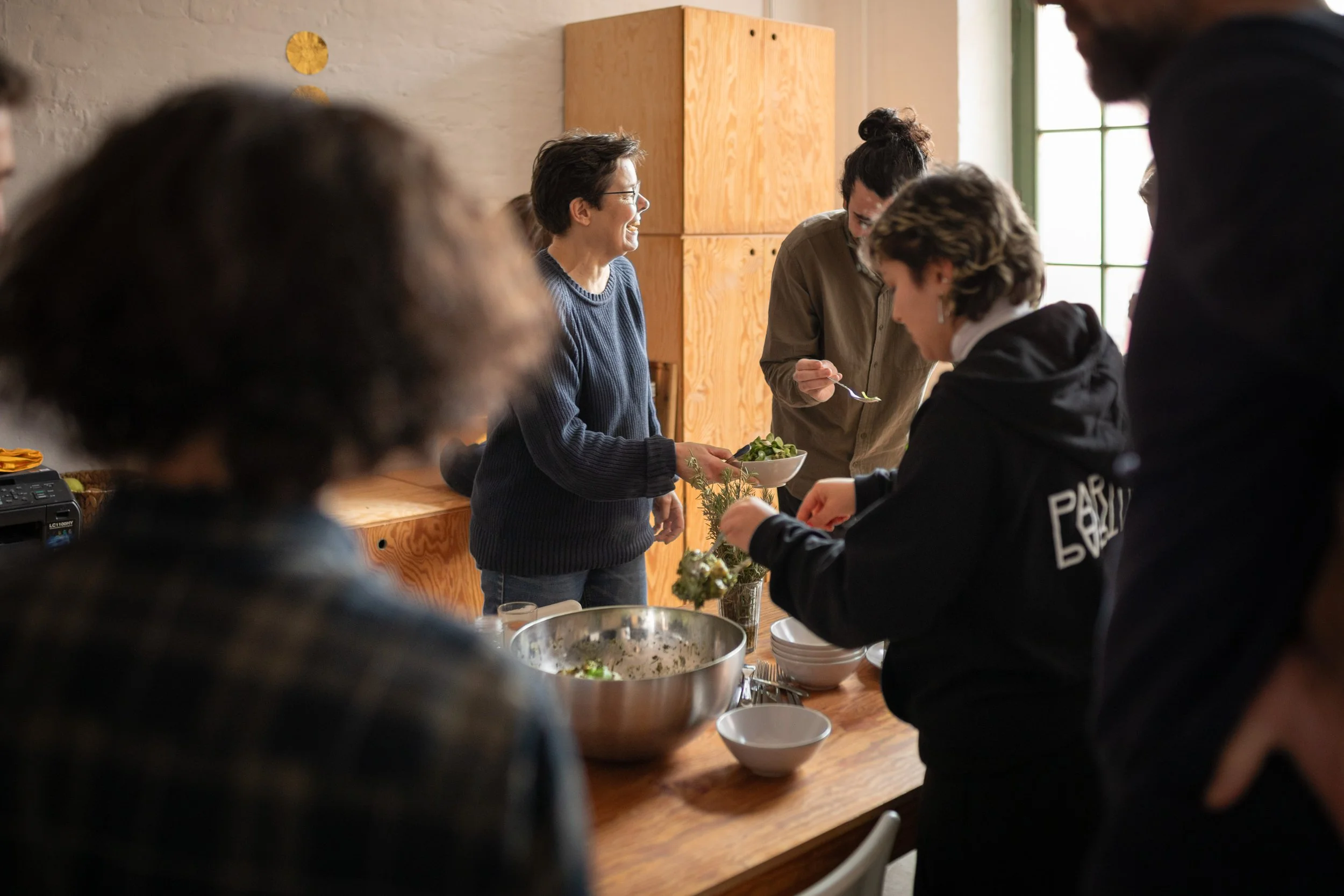 Menschen beim Kochen in einer modernen Küche mit Holzschränken und großen Fenstern, eine Person lächelt beim Servieren eines Salats.