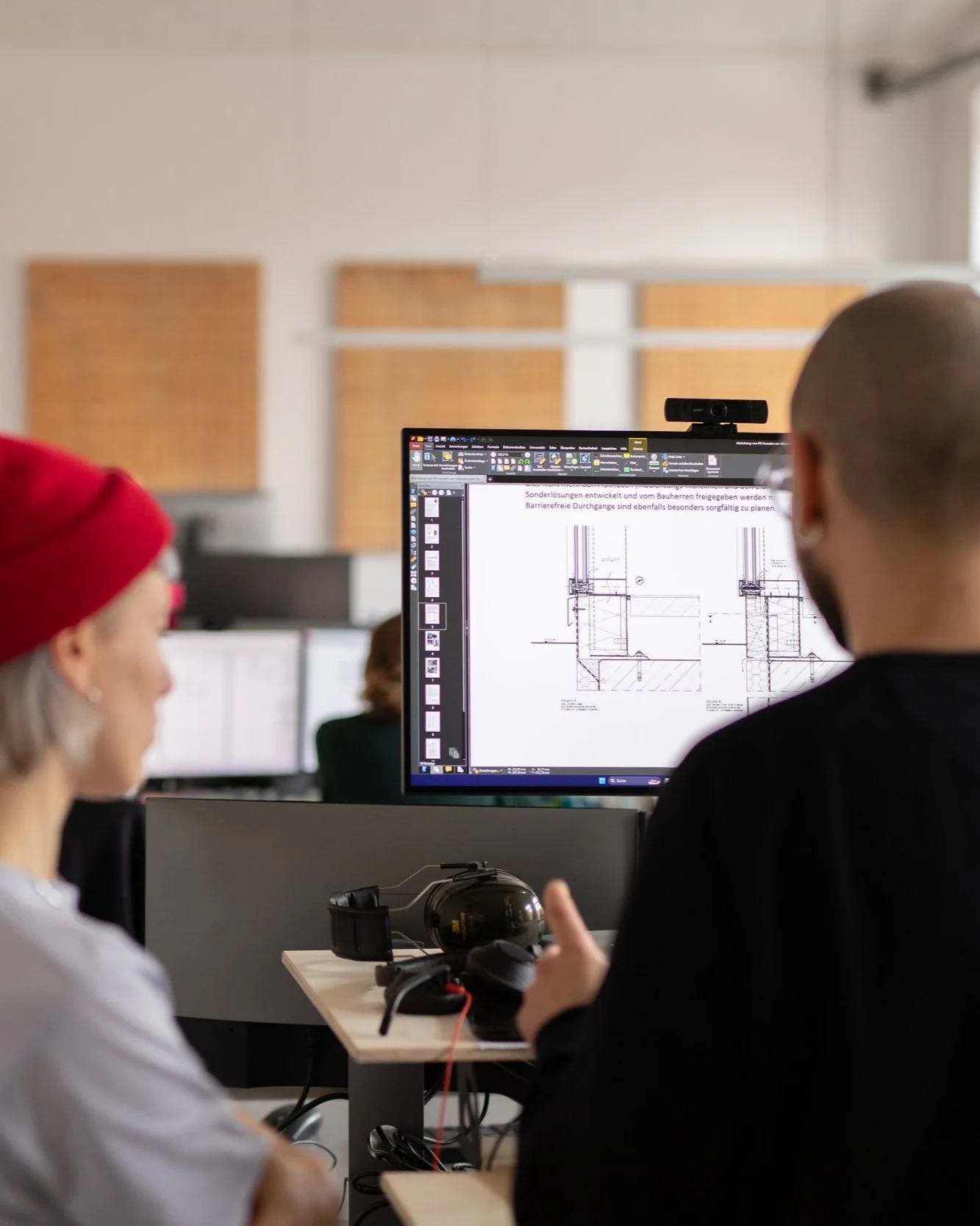 Zwei Menschen in einem Büro, die an Computern arbeiten. Auf dem Bildschirm eines Monitors sind technische Baupläne sichtbar.