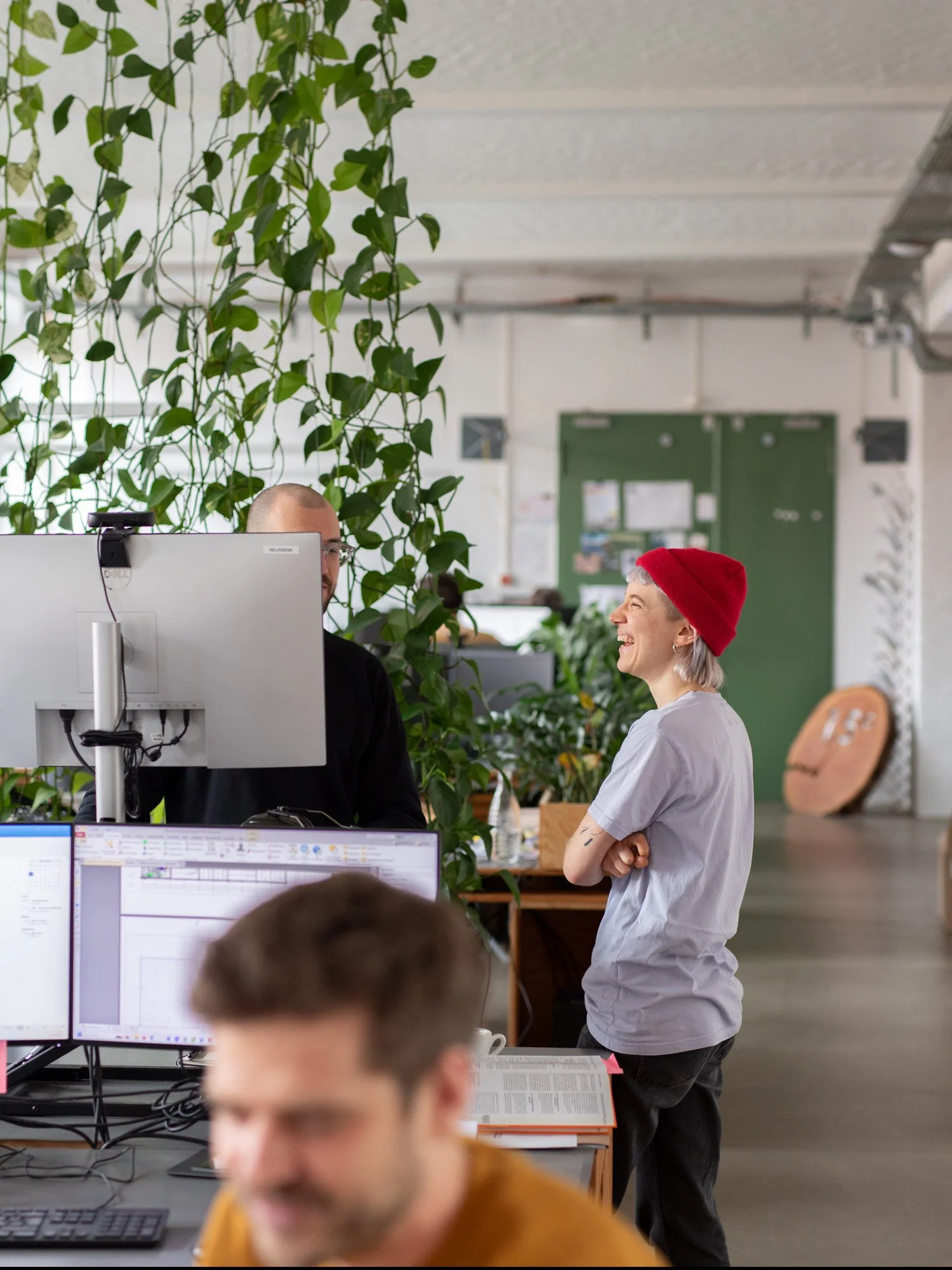 Zwei Personen, ein Mann mit Brille und Glatze, und eine Frau mit kurzen grauen Haaren und roter Mütze, lachen und sprechen in einem Büro. Im Vordergrund sind Monitore und Tastaturen sichtbar, im Hintergrund befinden sich Pflanzen und Büroeinrichtung.