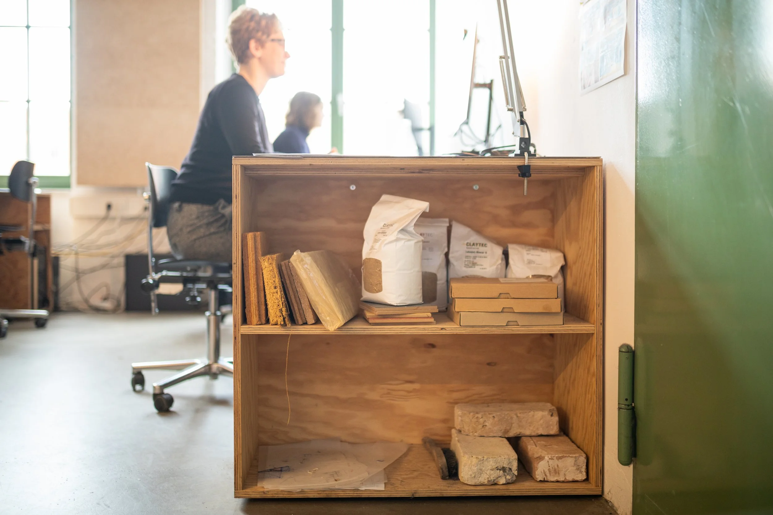 Ein Regal mit Baumaterialien und Verpackungen, in einem hell erleuchteten Raum, im Hintergrund zwei Personen an Computern.