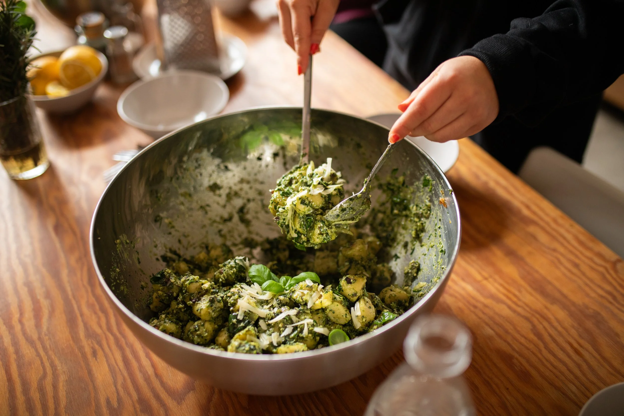 Person hebt einen grünen Kartoffelsalat mit Käse und frischen Kräutern in eine Schüssel. Im Hintergrund sind Zitronenscheiben, ein Glas Wasser und andere Küchenutensilien sichtbar.