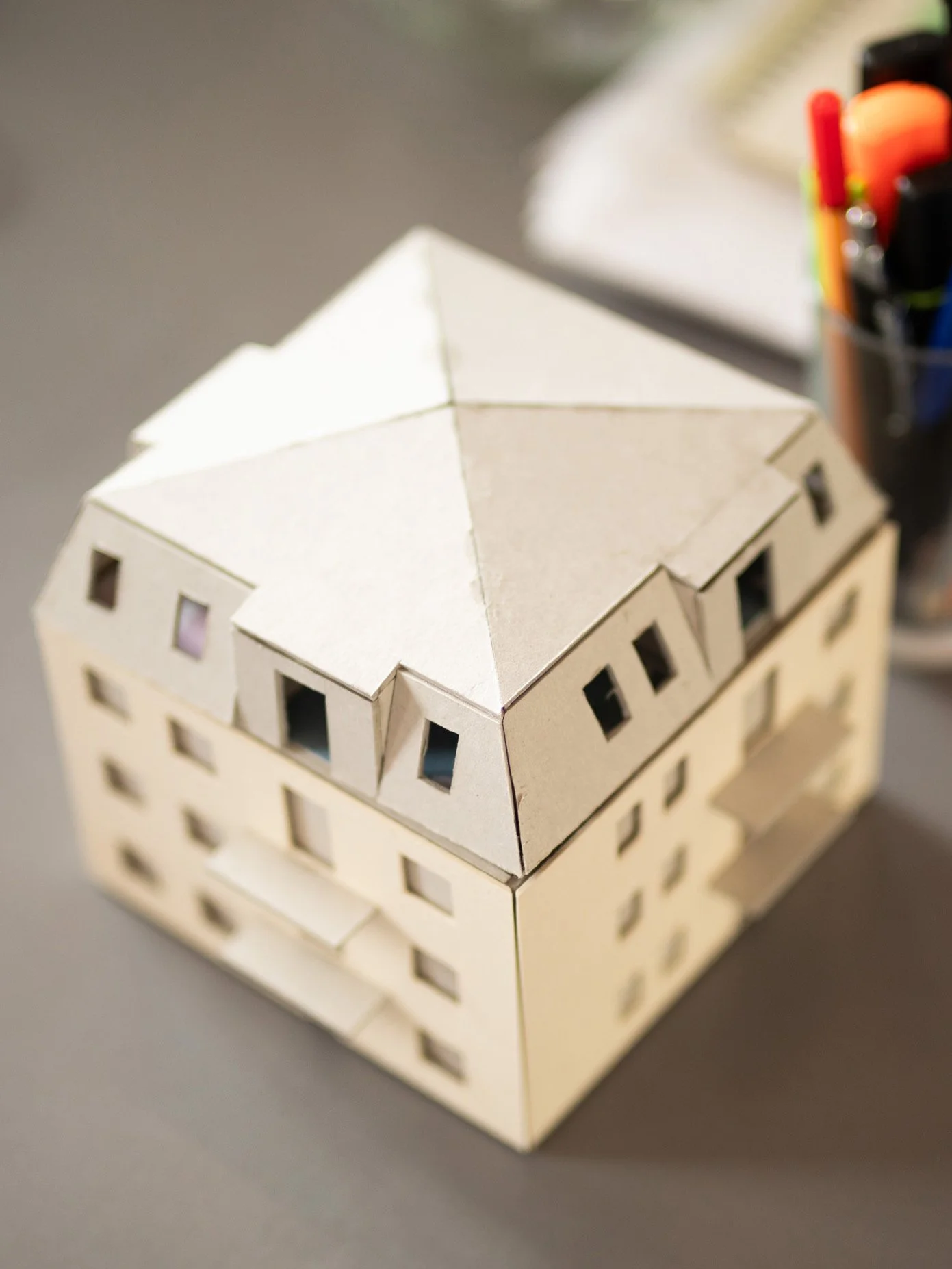 Ein Modellhaus aus Holz mit künstlichem Dach, daneben Stifte und Marker auf einem Tisch.