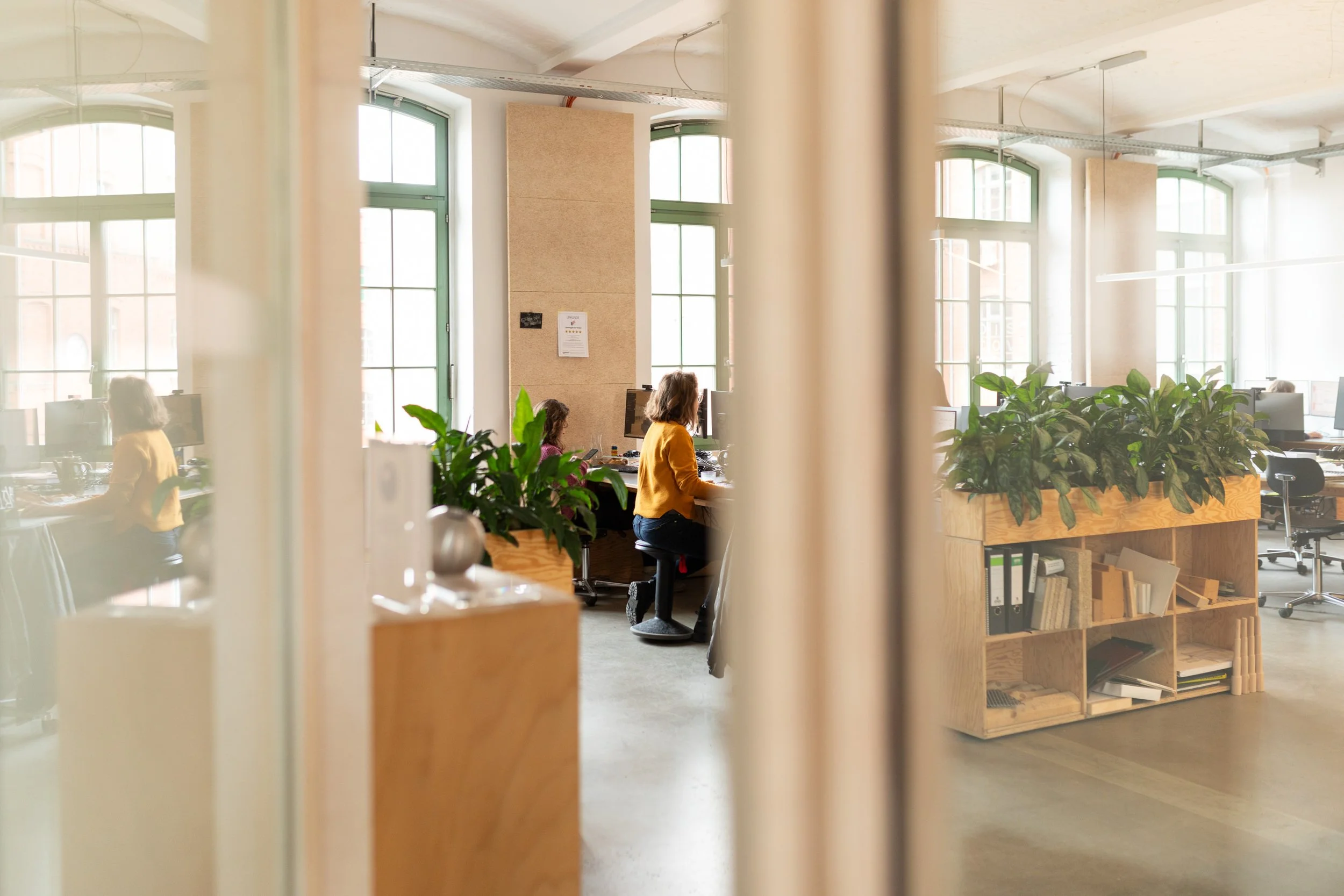 Innenansicht eines modernen Büros mit großen Fenstern, mehreren Personen an Computern und einer Holzwand mit Pflanzen im Vordergrund.