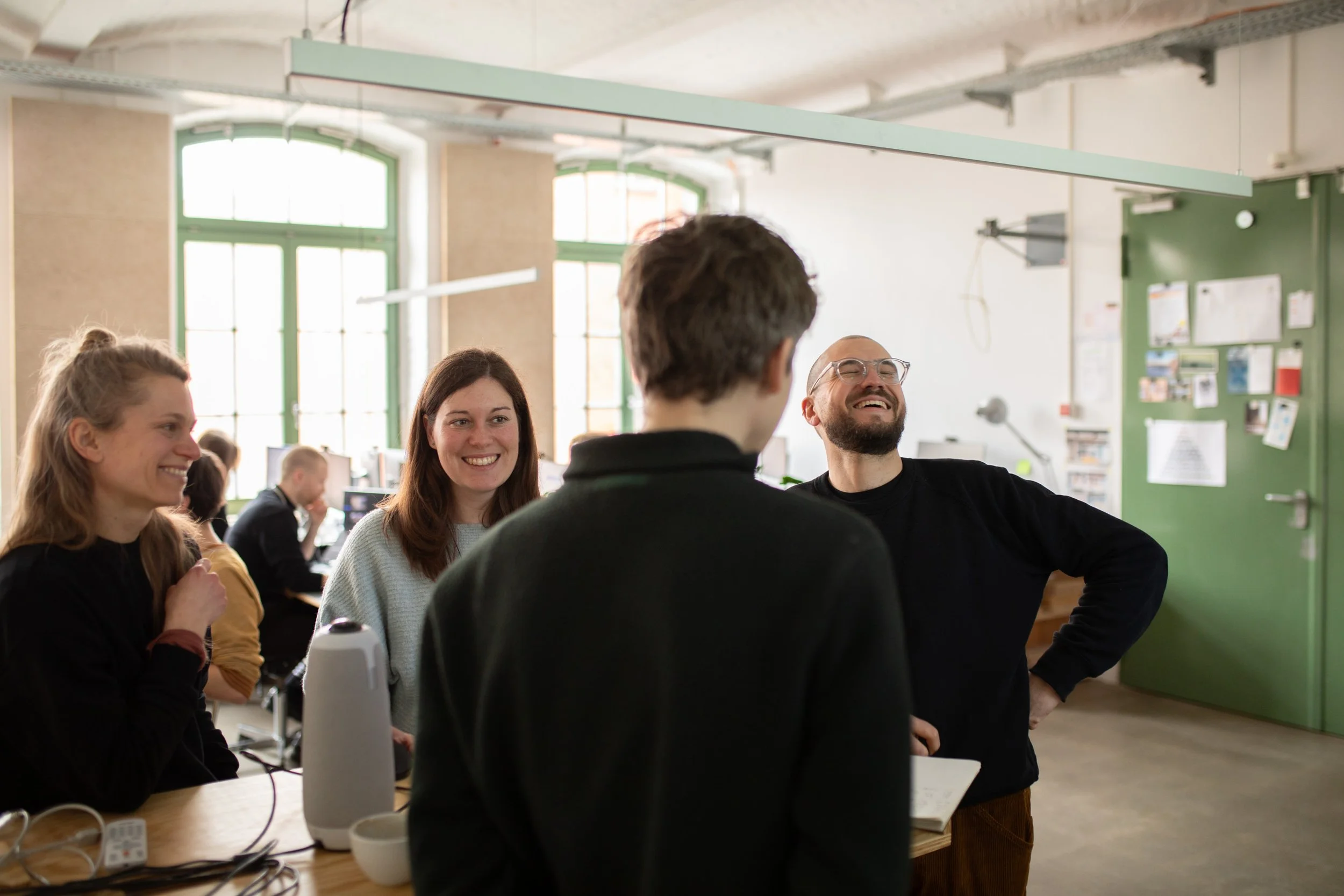 Gruppe von Menschen, die in einem hellen Raum zusammen lachen und sprechen. Manche sitzen an einem Tisch, auf dem technische Geräte und Utensilien liegen.