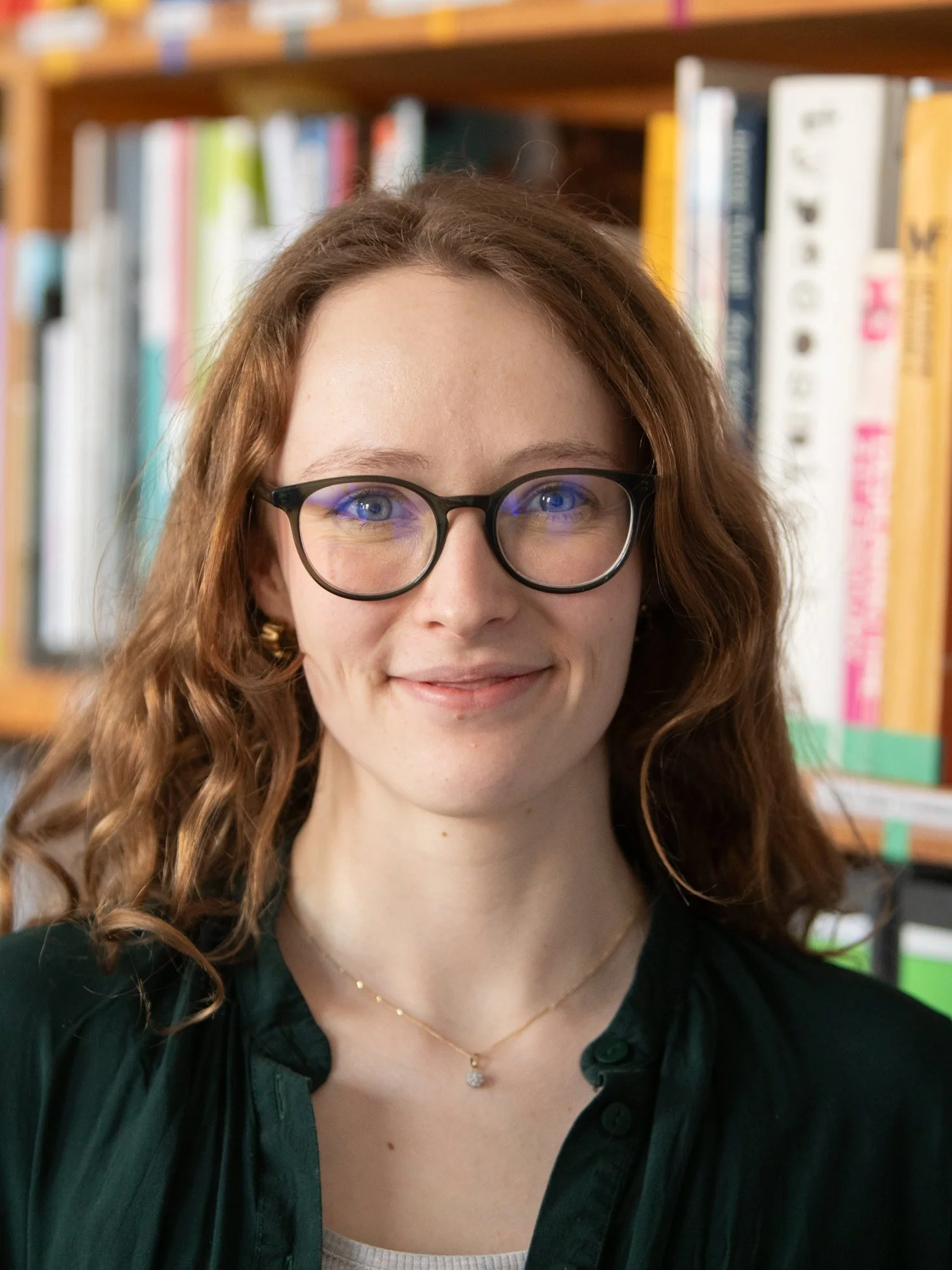 Porträt einer Frau mit lang, lockigem, braunem Haar, schwarzer Brille, Lächeln, im Hintergrund Bücherregal.