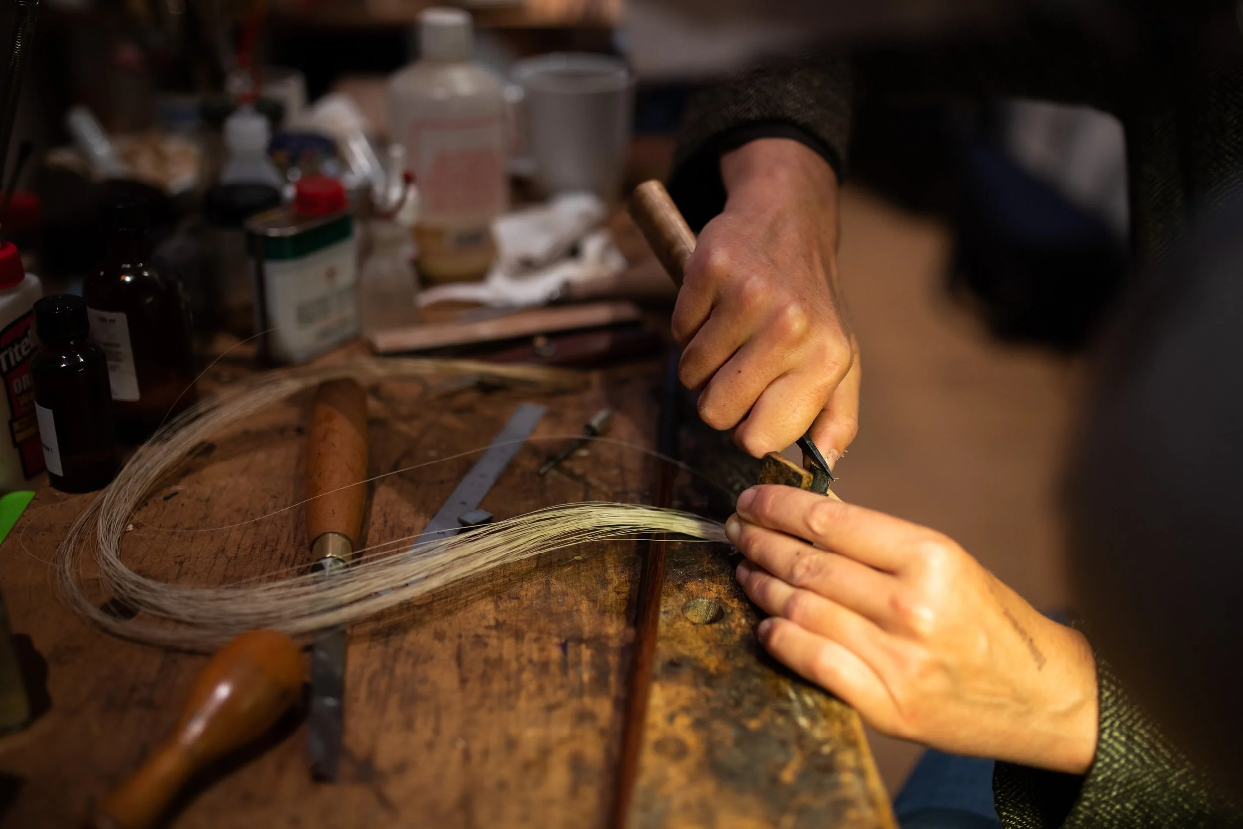 Handwerkliche Arbeit mit einem Messer an einem Holzstück, im Hintergrund verschiedene Flaschen und Behälter auf einem Tisch.