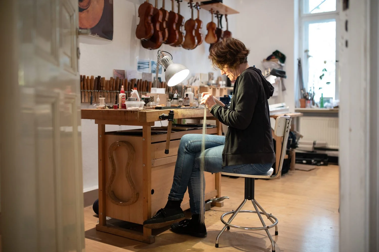 Junge Frau arbeitet in einer Werkstatt mit Geigen, sitzt auf einem Hocker vor einem Holztisch mit Werkzeugen und Materialien, hinter ihr sind mehrere Geigen an der Wand aufgehängt.
