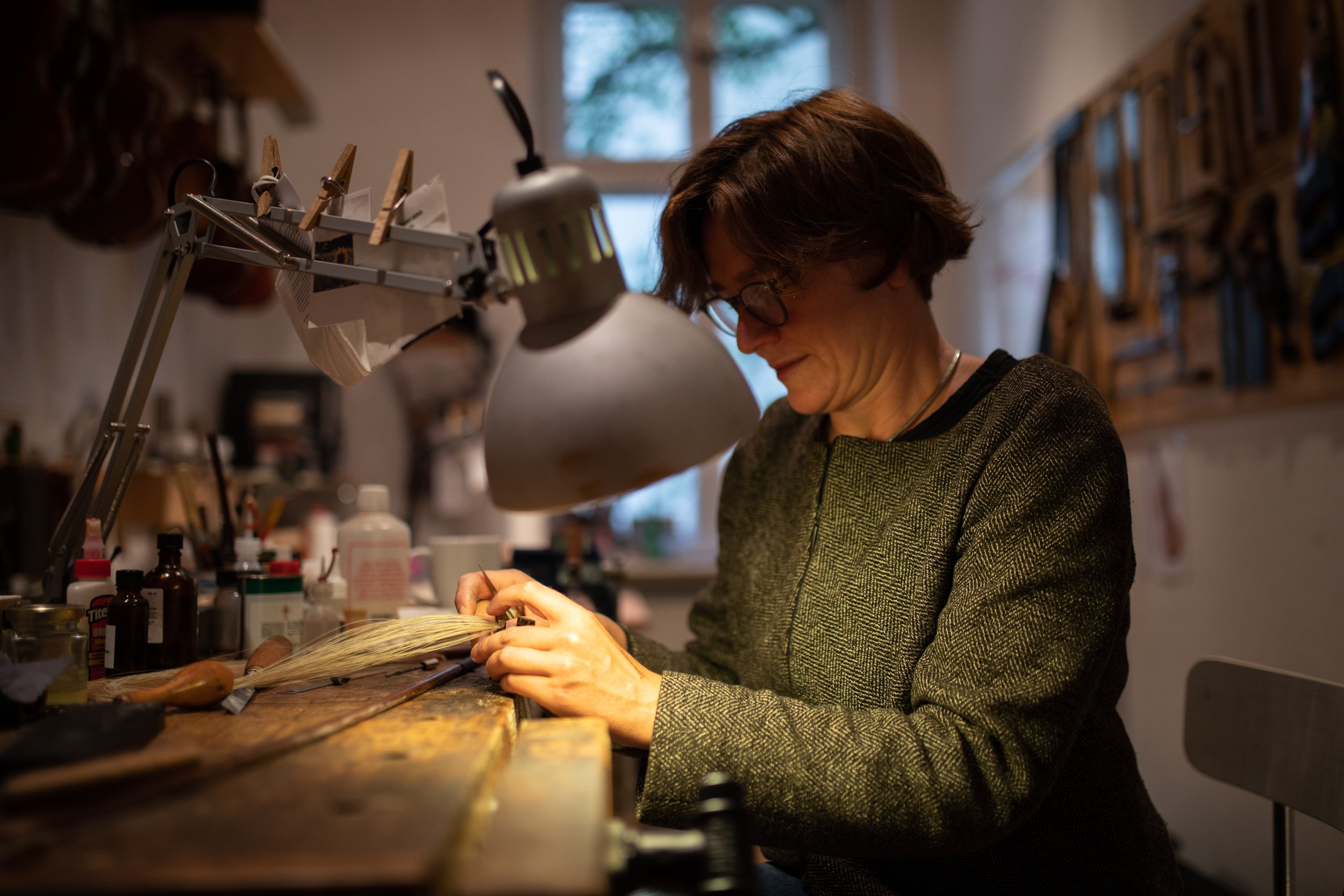 Eine Frau mit kurzen braunen Haaren und Brille arbeitet an einer Werkbank in einem Atelier, umgeben von Werkzeugen und Materialien, vermutlich beim Anfertigen oder Reparieren eines Gegenstands.