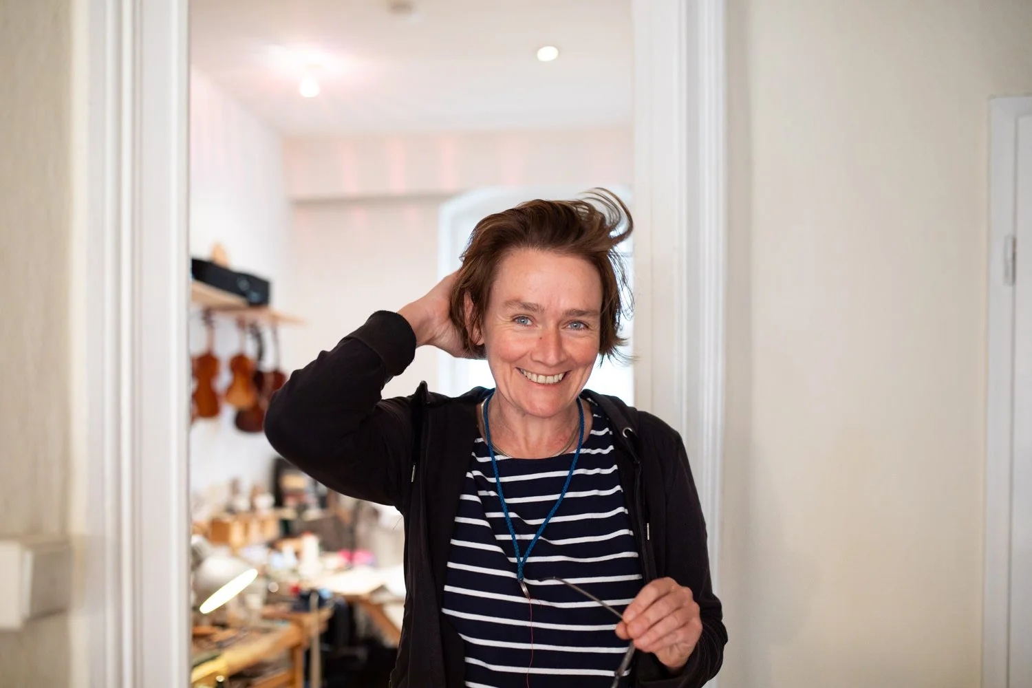 Eine lachende Frau mit braunen Haaren im Türrahmen, gehaltene Brille in der Hand, im Hintergrund ein Raum mit Musikinstrumenten und Computer.