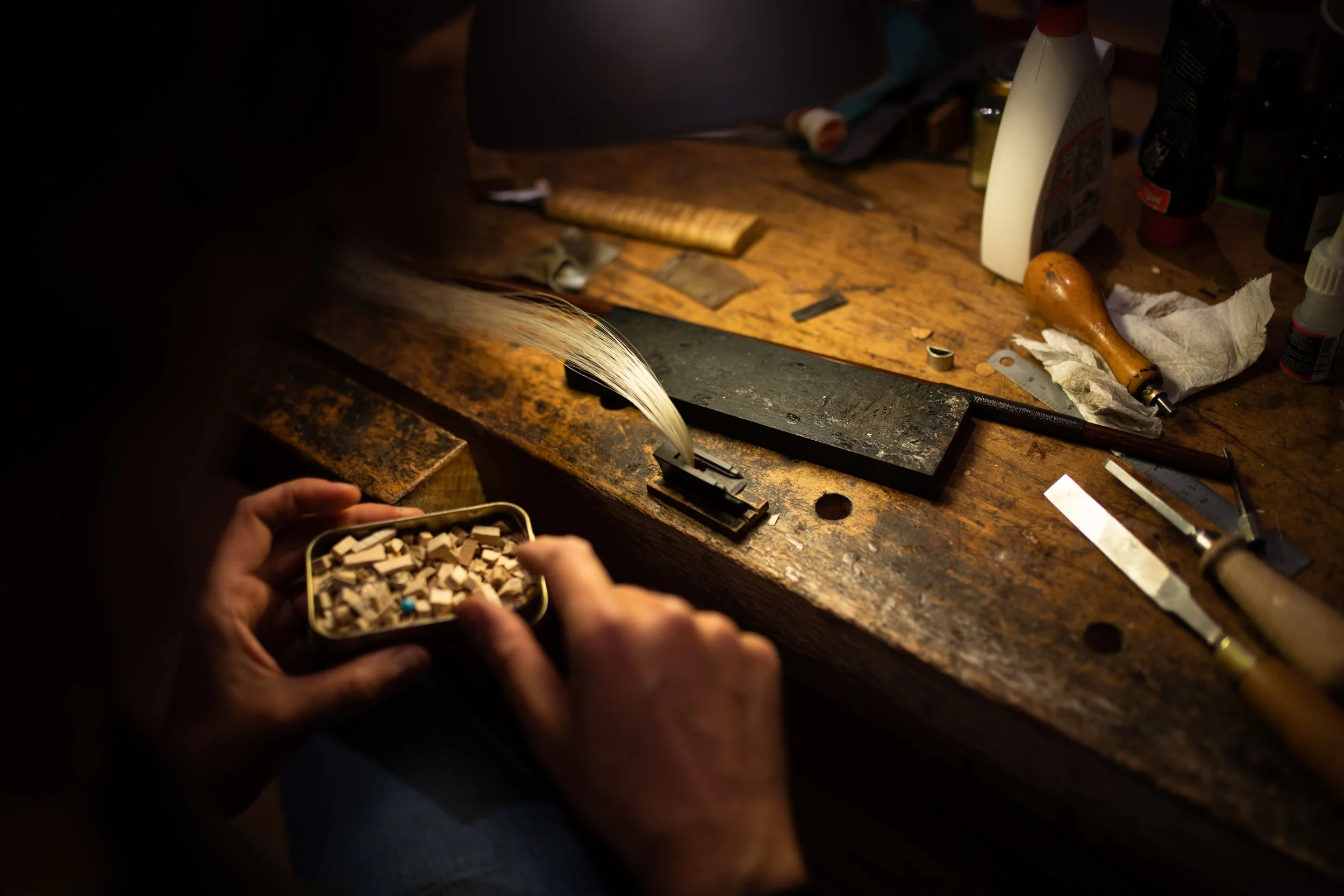 Personen beim Herstellen eines Musikinstruments, vermutlich eines Violins. Auf dem Tisch liegen Werkzeuge, Holzstäbchen und Kleinteile, die für die Handwerkskunst verwendet werden.