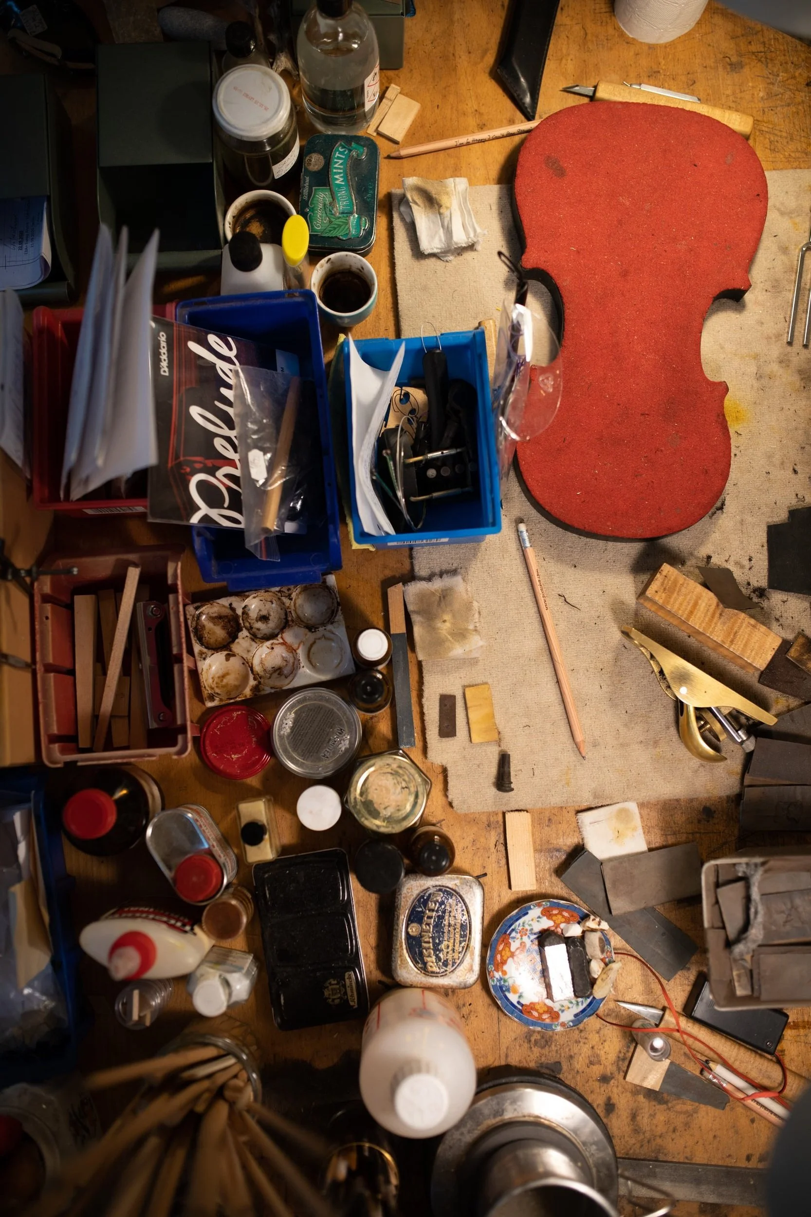 Ein unordentischer Arbeitstisch mit verschiedenen Gegenständen, darunter Flaschen, Dosen, Werkzeuge, Papiere, eine rote Stoffmatte, eine Schale mit Eiern und diverses Werkzeug. Es sieht nach einer Bastel- oder Reparaturwerkstatt aus.