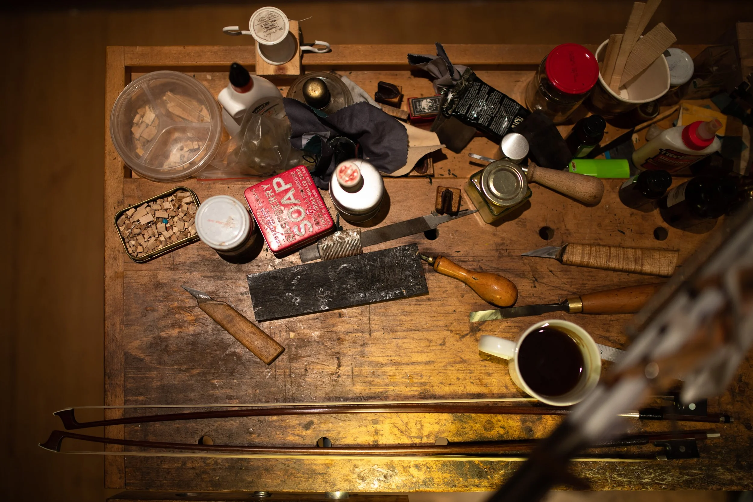 Ein unordentischer Holztisch mit verschiedenen Kerben, Fläschchen, Gläsern, Schrauben, einem Messer, einer Klinge, einer Zange, Kaugummi, Papier, einer Flasche, und einem Becher mit schwarzem Kaffee.