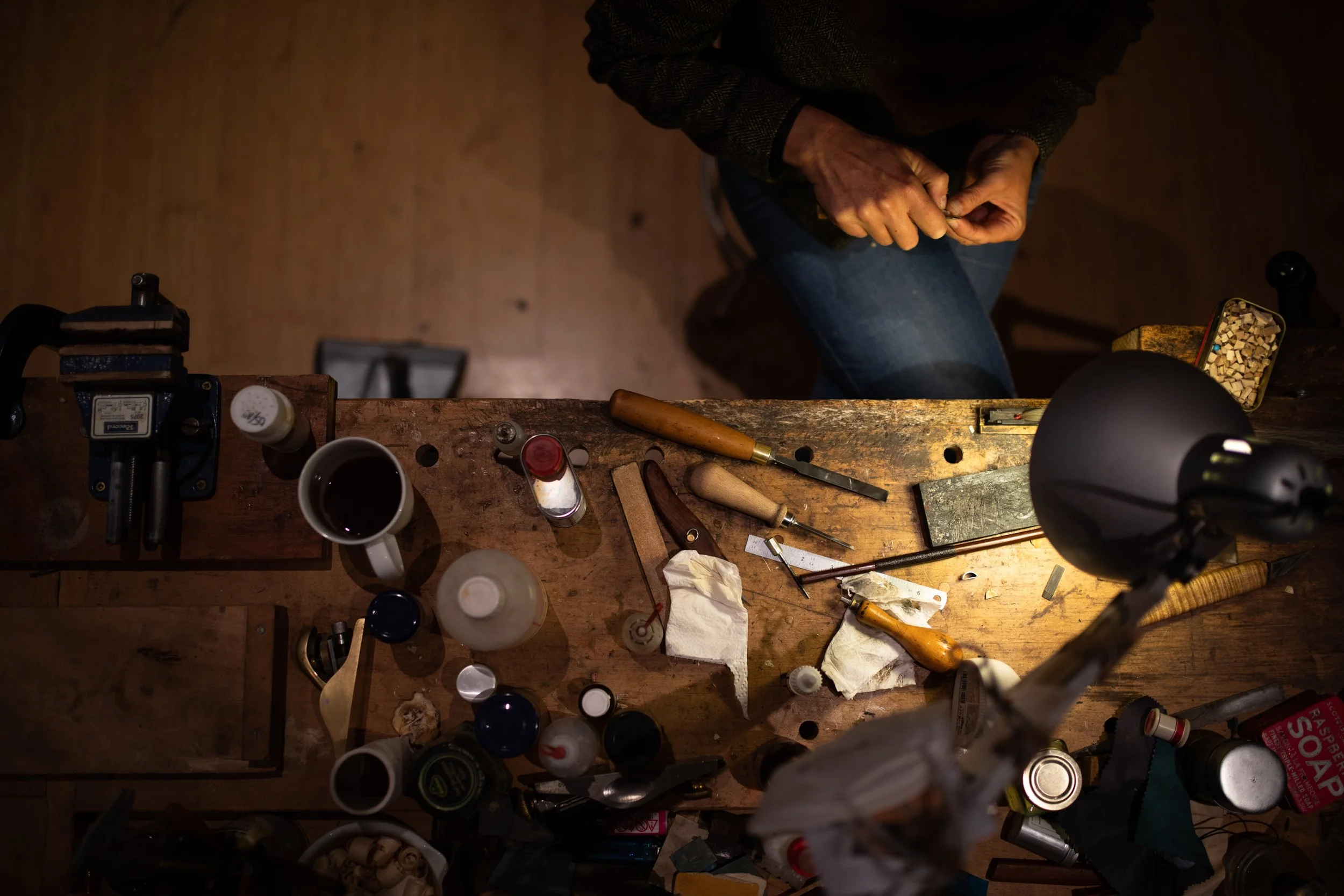 Ein Oberkörper einer Person, die an einem Holzarbeitsbereich arbeitet, umgeben von Werkzeugen, Farbflaschen und Werkstücken in einer Werkstatt.