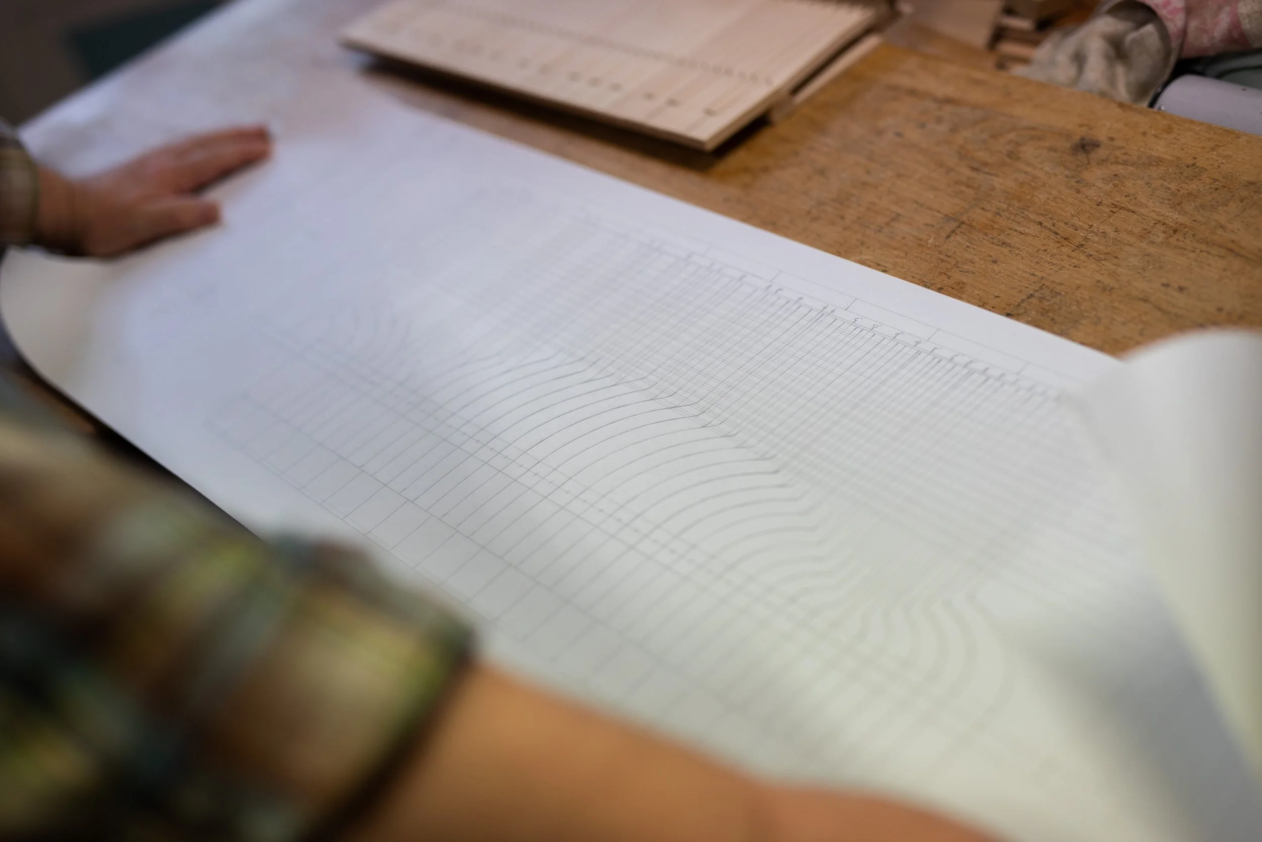 Blick auf eine Hand, die auf einem Holzarbeitstisch ein aufgeschlagenes Buch mit graphischer Darstellung und ein weiteres Buch mit Tabellen und Texten betrachtet.