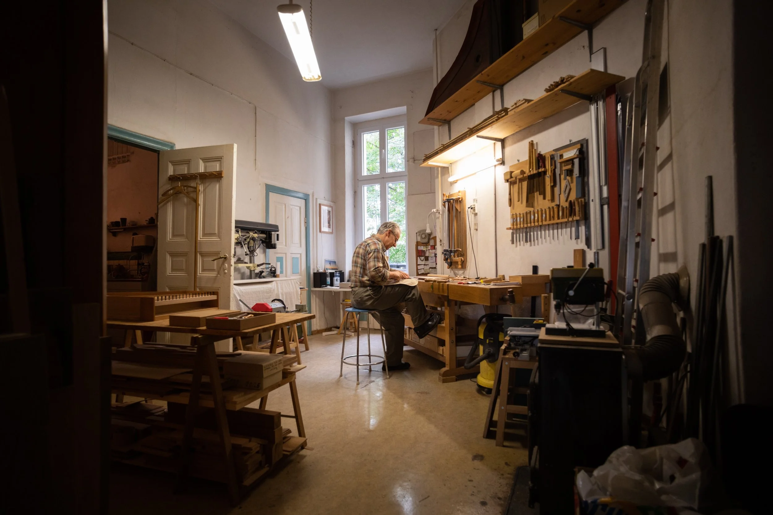 Ein älterer Mann arbeitet in einer Werkstatt an einem Holzprojekt. Die Werkstatt hat hohe Fenster und Werkbänke mit Werkzeugen, Regale und eine Leiter.