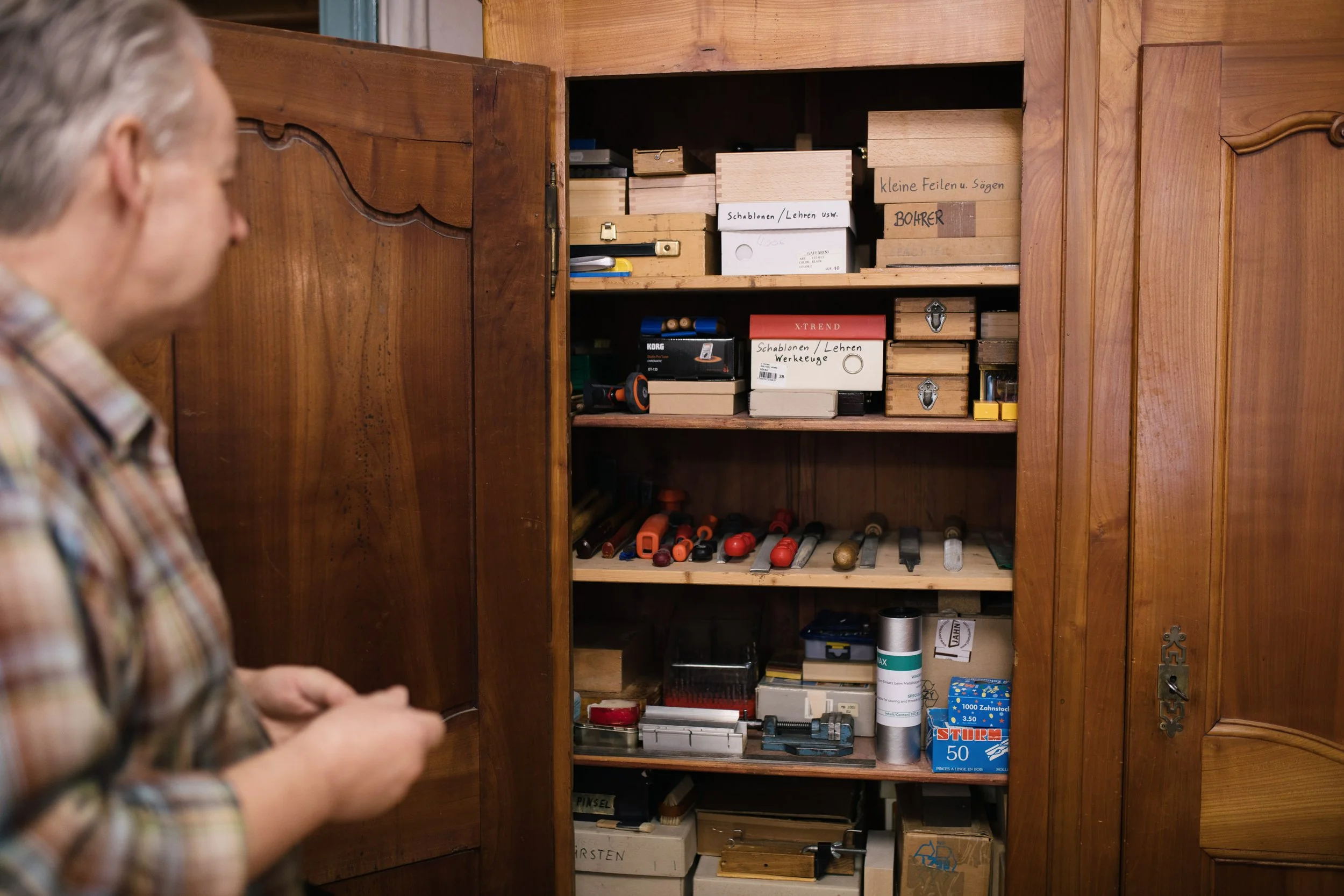 Ein Mann betrachtet einen Holzschrank mit verschiedenen Werkzeugen und Materialien, darunter Schraubenzieher, Holzstücke, eine Dose und kleine Holzteile, beschriftet mit handgeschriebenen Labels.