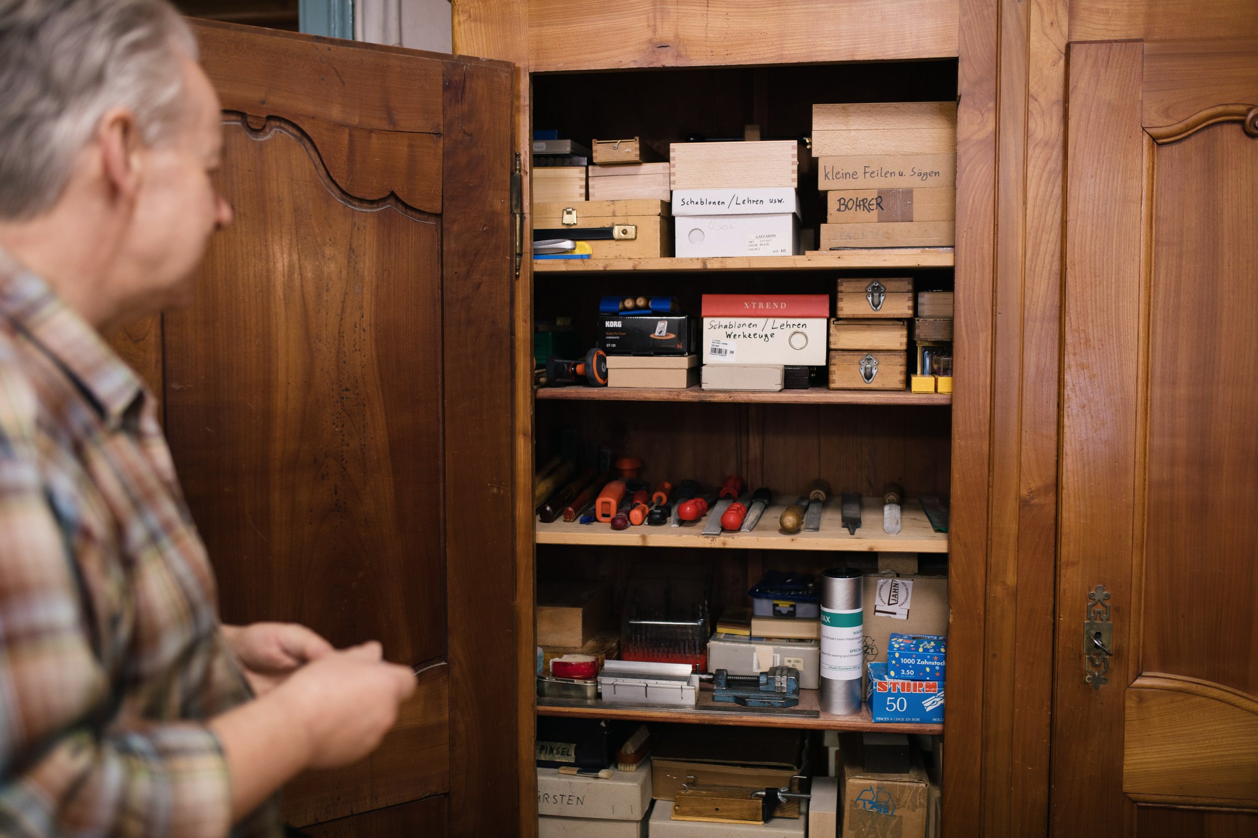 Ein Mann betrachtet einen Holzschrank mit verschiedenen Werkzeugen und Materialien, darunter Schraubenzieher, Holzstücke, eine Dose und kleine Holzteile, beschriftet mit handgeschriebenen Labels.