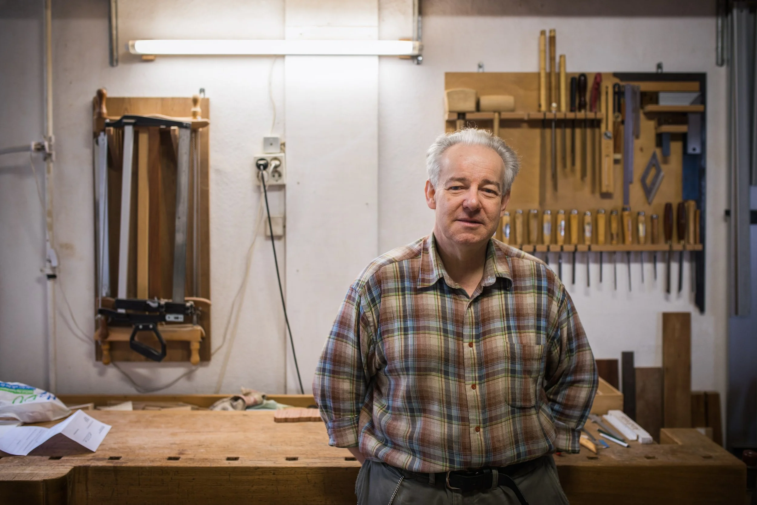 Ein älterer Mann steht in einer Werkstatt vor einer Werkbank. Im Hintergrund sind Spachtel, Sägen und andere Handwerkzeuge an einer Holzwand befestigt.