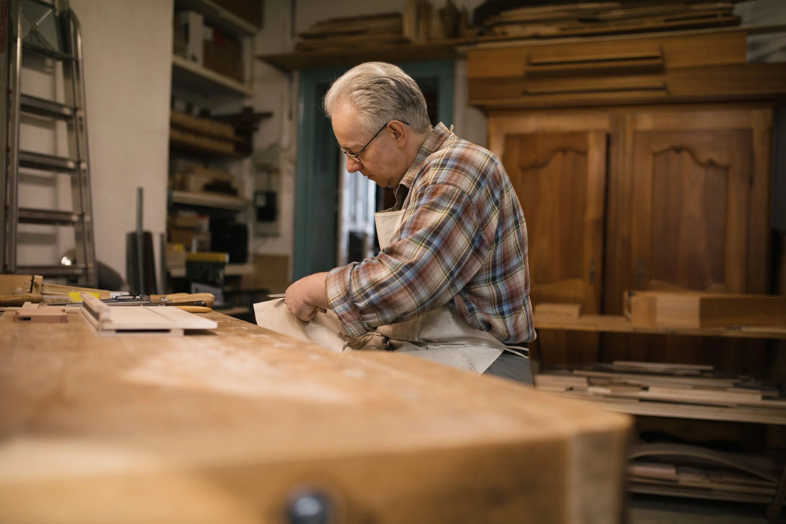 Älterer Mann in einer Werkstatt, der an einem Werkstück arbeitet, umgeben von Holzmaterialien und Werkzeugen.