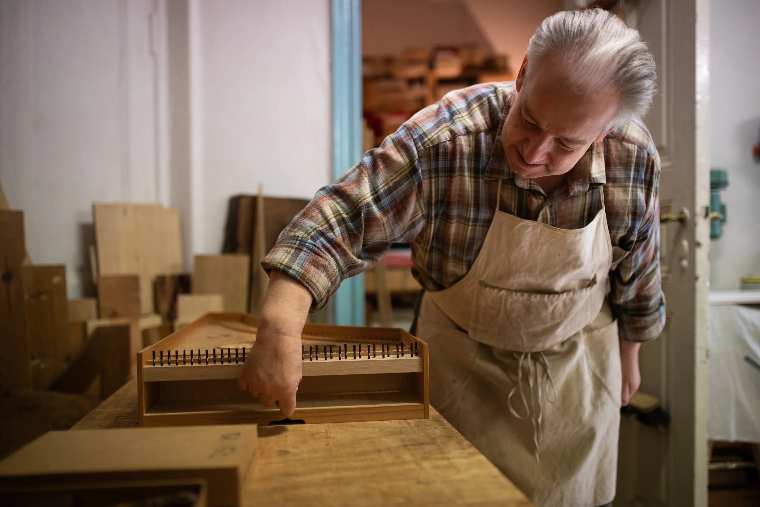 Älterer Mann in kariertem Hemd und Schürze arbeitet in einer Holzwerkstatt an einem kleinen Holzstück.