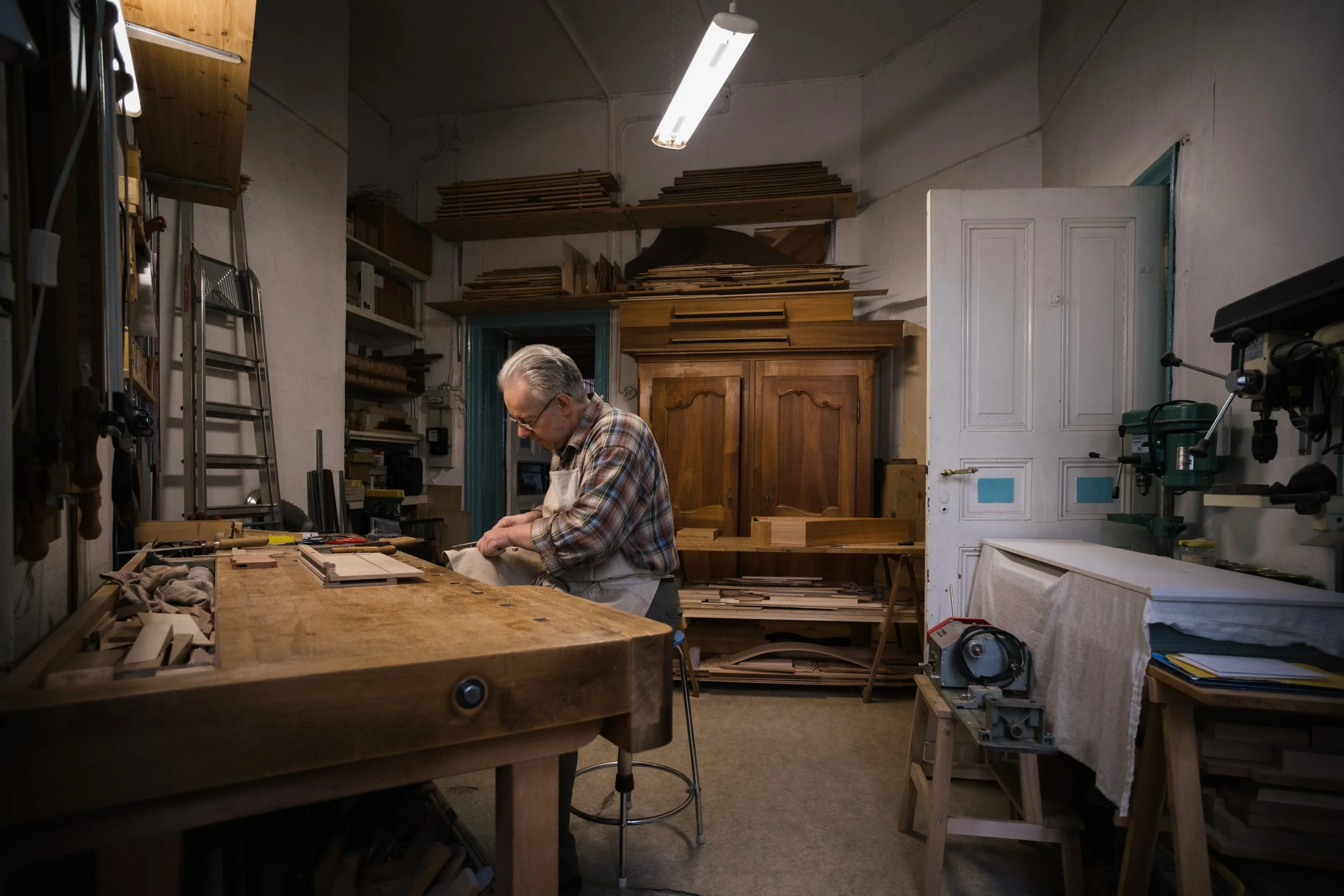 Ein älterer Mann arbeitet in einer Holzwerkstatt, umgeben von Werkzeugen, Holz und Möbelstücken, mit einer Halogenlampe an der Decke.