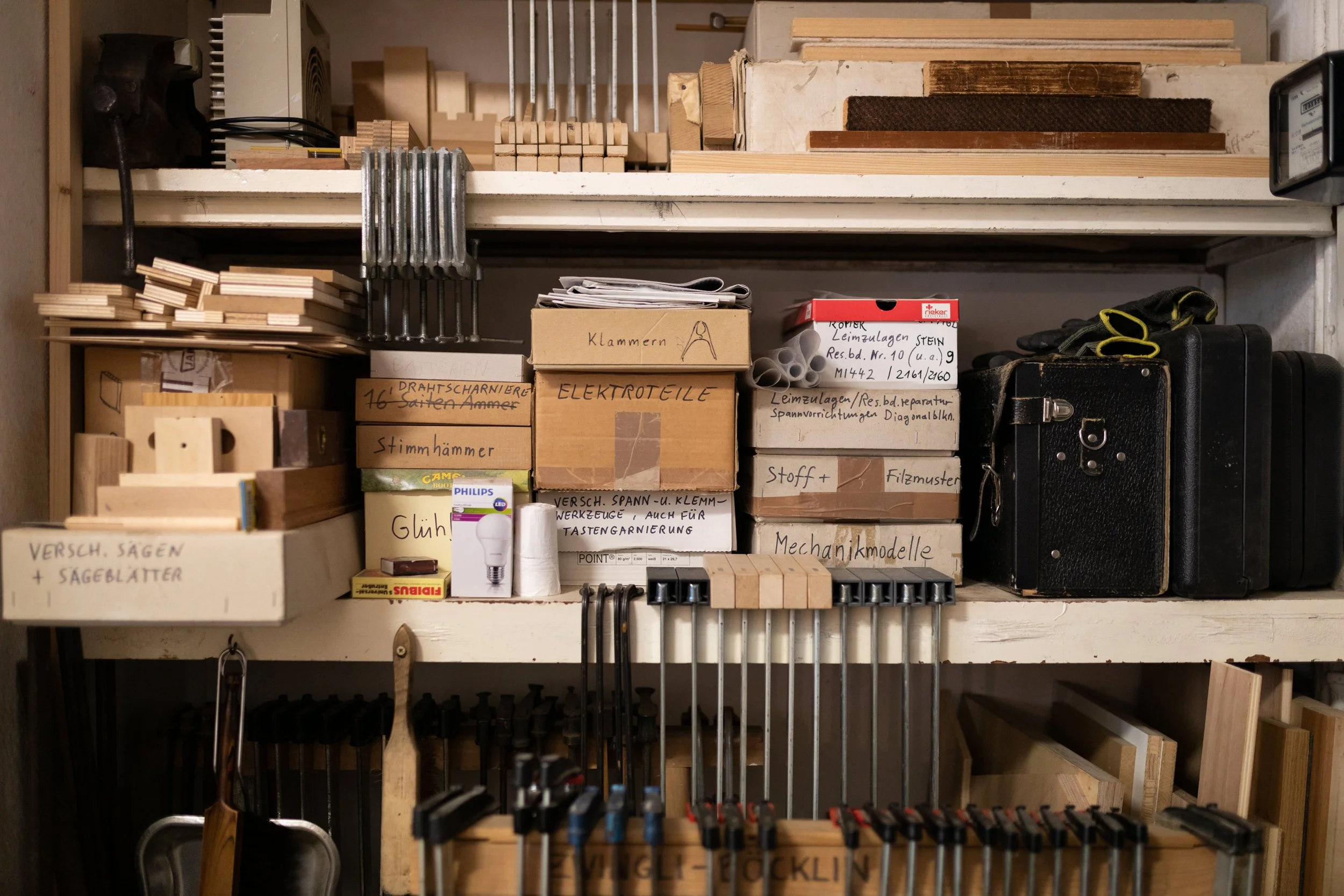 Werkstattregal mit verschiedenen handwerklichen Materialien und Werkzeugen. Darunter befinden sich Holzstücke, Schrauben, Boxen mit Schrauben, Kabel, Hammer, und elektrische Komponenten.