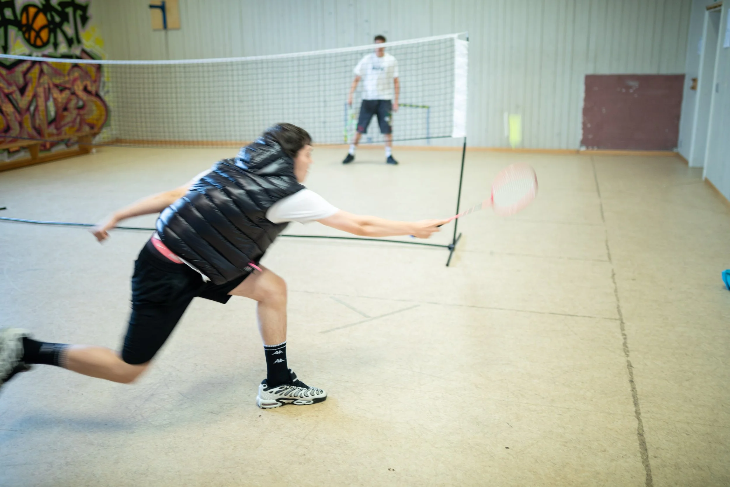 Zwei Jugendliche spielen Badminton in einer Turnhalle. Der Junge im Vordergrund ist in Bewegung, während im Hintergrund ein anderes Kind vor einem Netz steht.