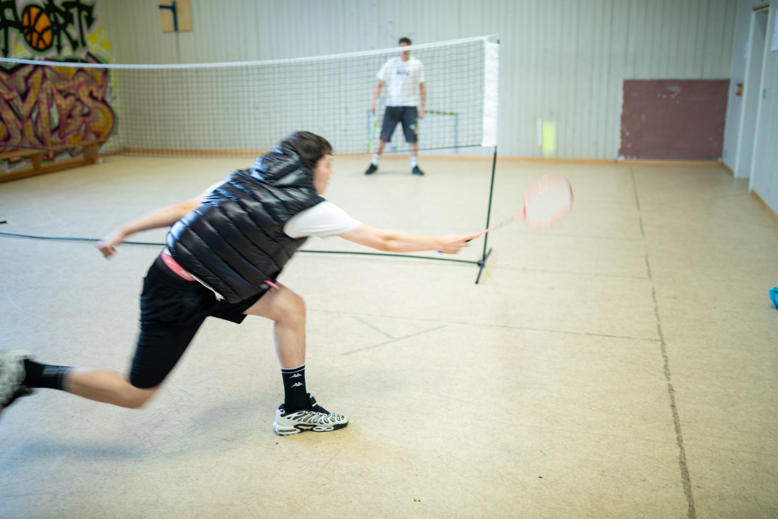 Zwei Jugendliche spielen Badminton in einer Turnhalle. Der Junge im Vordergrund ist in Bewegung, während im Hintergrund ein anderes Kind vor einem Netz steht.