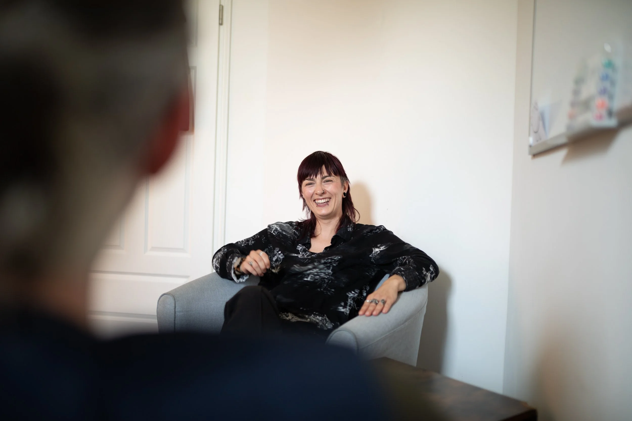 Eine lachende Frau sitzt auf einem Sofa in einem Raum, während jemand anderes im Vordergrund unscharf ist.