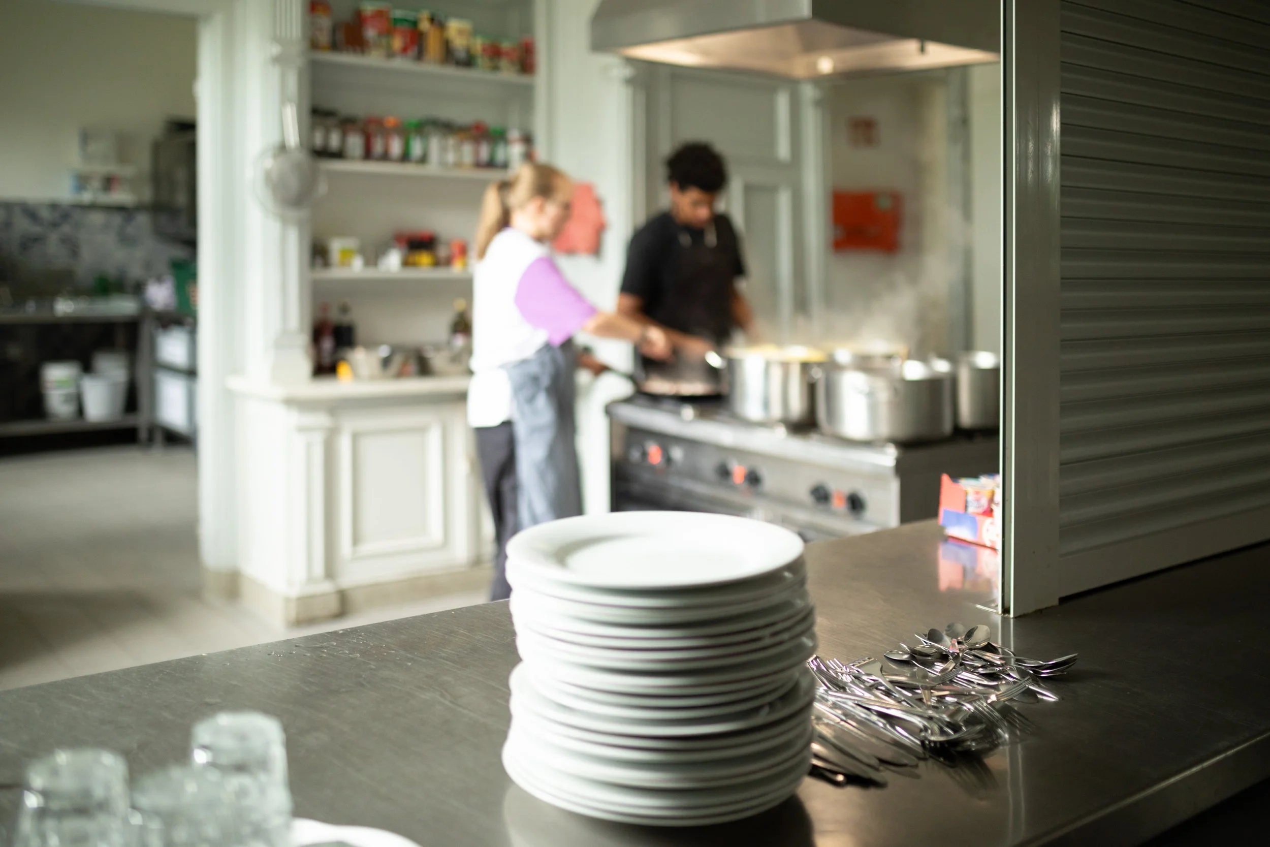 Menschen beim Kochen in einer modernen Küche, im Vordergrund gibt es stapelweise weiße Teller, Löffel und Gabeln.