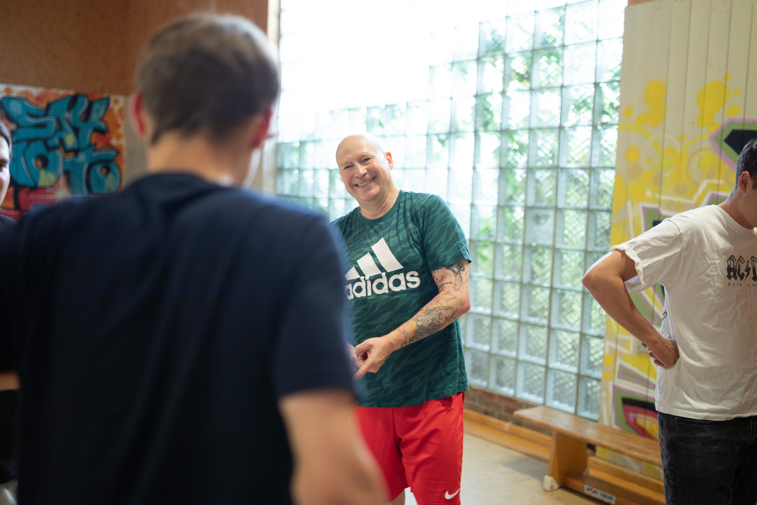Mehrere Personen, darunter ein Mann mit einem grünen Adidas-T-Shirt, stehen in einem Raum mit großen Fenstern und Graffiti an den Wänden, während sie sich unterhalten.