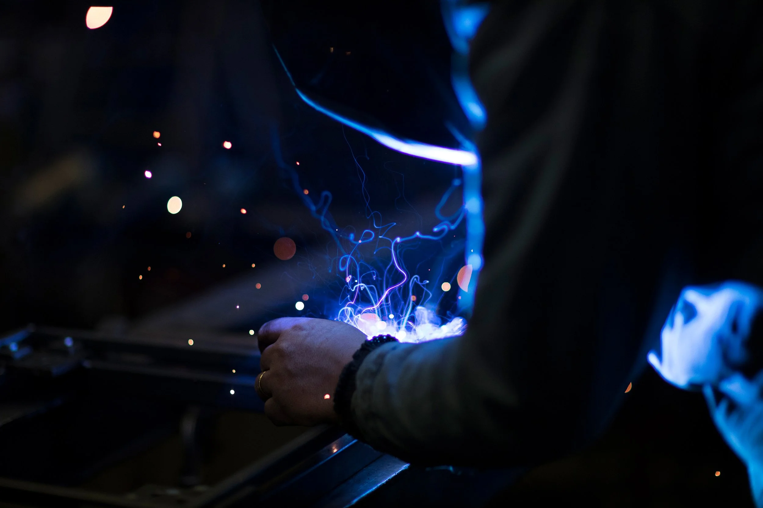 Person schweißt Metall, blauer Lichtstrahl und Funken in dunklem Hintergrund sichtbar.