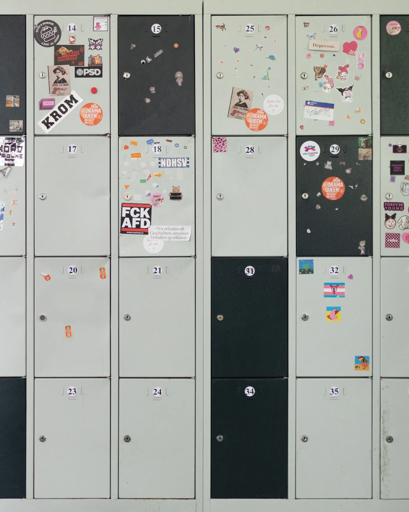 Eine Reihe von verschlossenen schwarzen und grauen Schließfächern mit Nummern und bunten Aufklebern, teilweise mit Sticker und Zetteln.