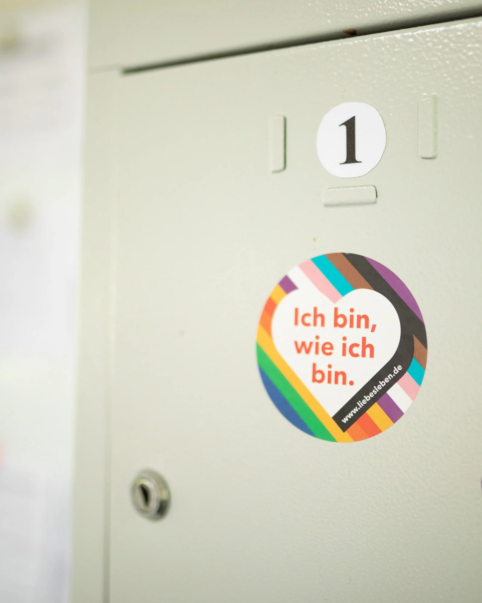 Auf einem weißen Kasten ist ein Aufkleber mit einem Regenbogenrahmen und einem Slogan in deutscher Sprache: "Ich bin, wie ich bin", außerdem eine Website-Adresse. Ein Türschloss ist sichtbar.