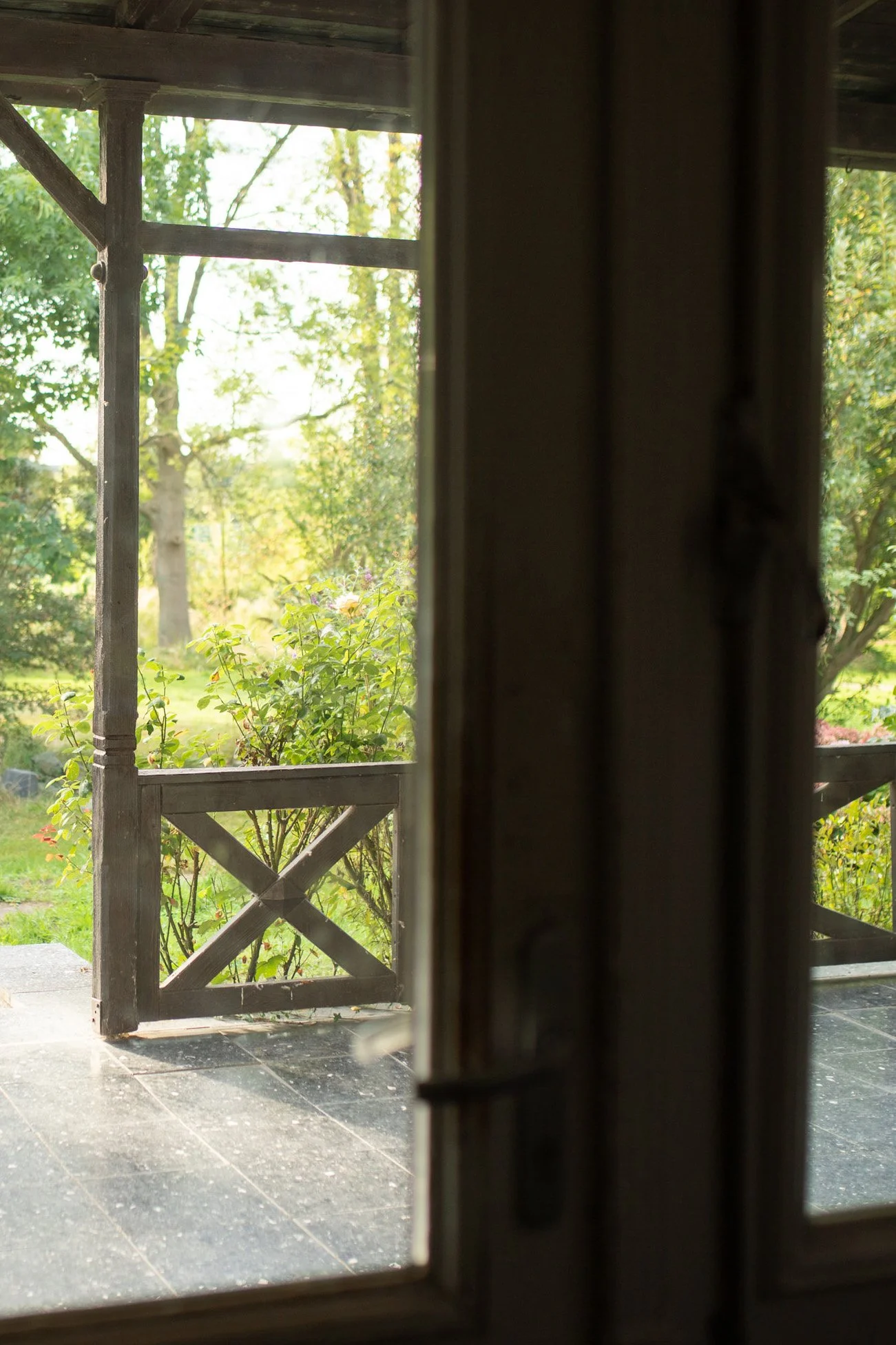 Blick durch ein offenes Fenster auf eine Veranda mit Holzgeländer, grünem Garten und Bäumen im Hintergrund.