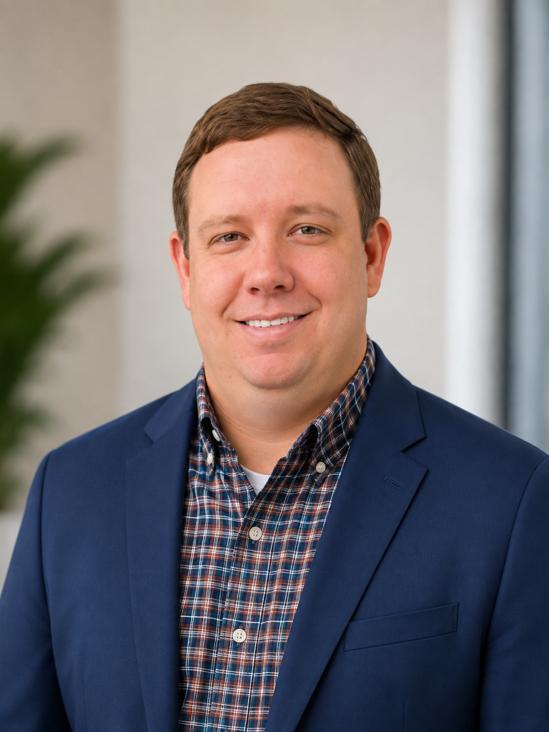 A man in a blue blazer and plaid shirt smiling for a portrait in an indoor setting.