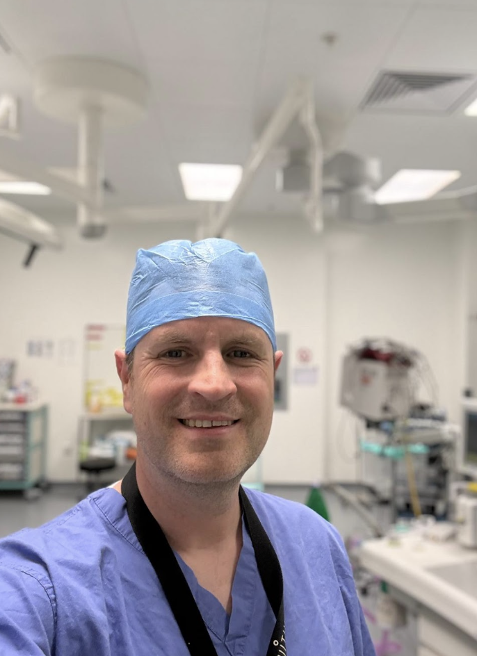 A smiling male healthcare professional in scrubs and a surgical cap in a hospital or medical facility.
