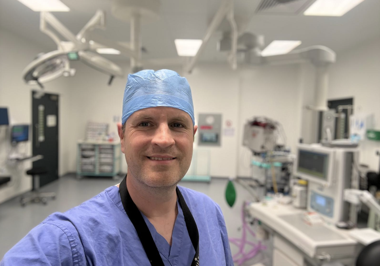 A male healthcare professional wearing blue scrubs, a surgical cap, and a lanyard, taking a selfie in a hospital operating room with medical equipment and monitors in the background.