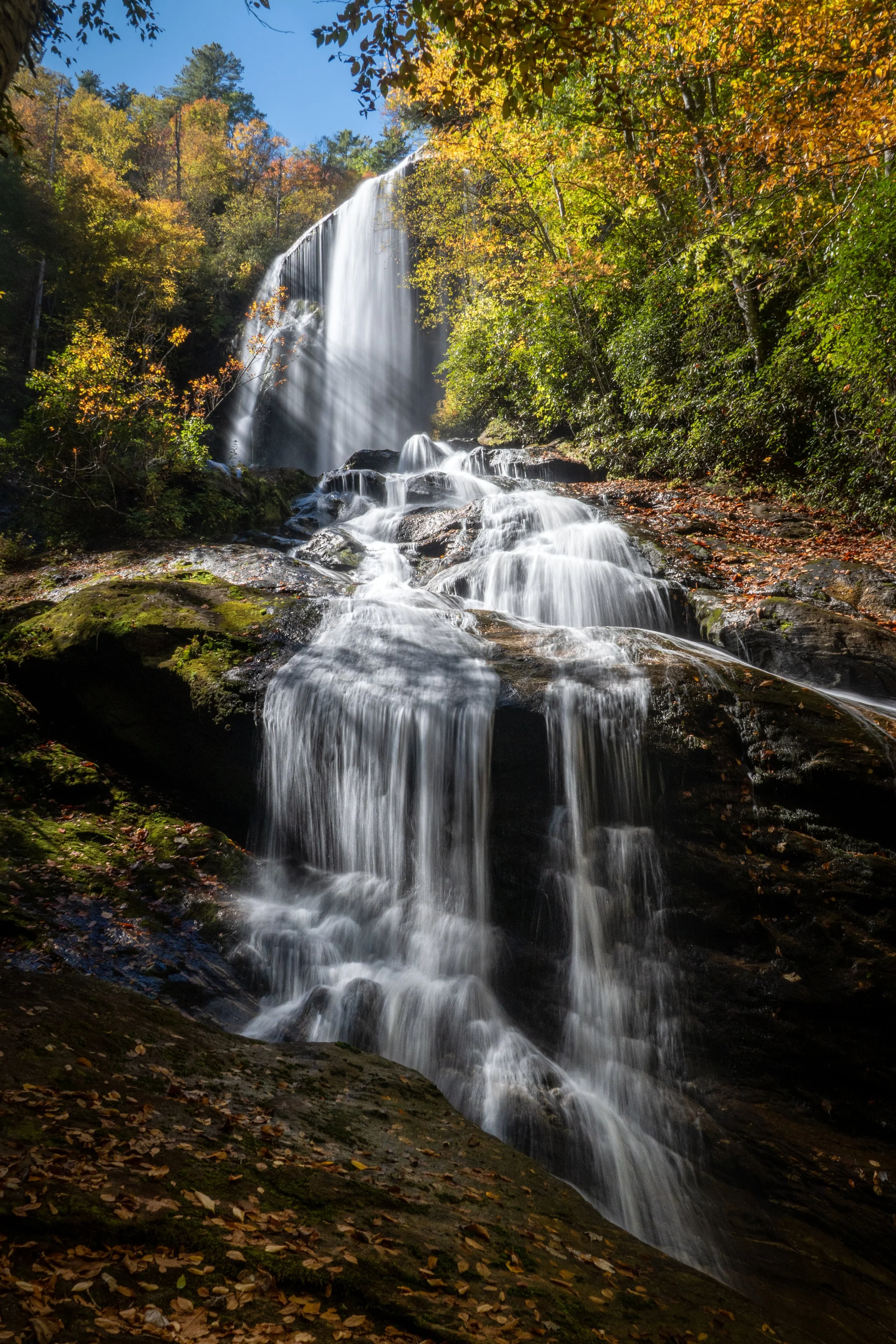 A cascade of waterfalls flowing down a rocky landscape surrounded by trees with fall foliage, bright blue sky in the background.