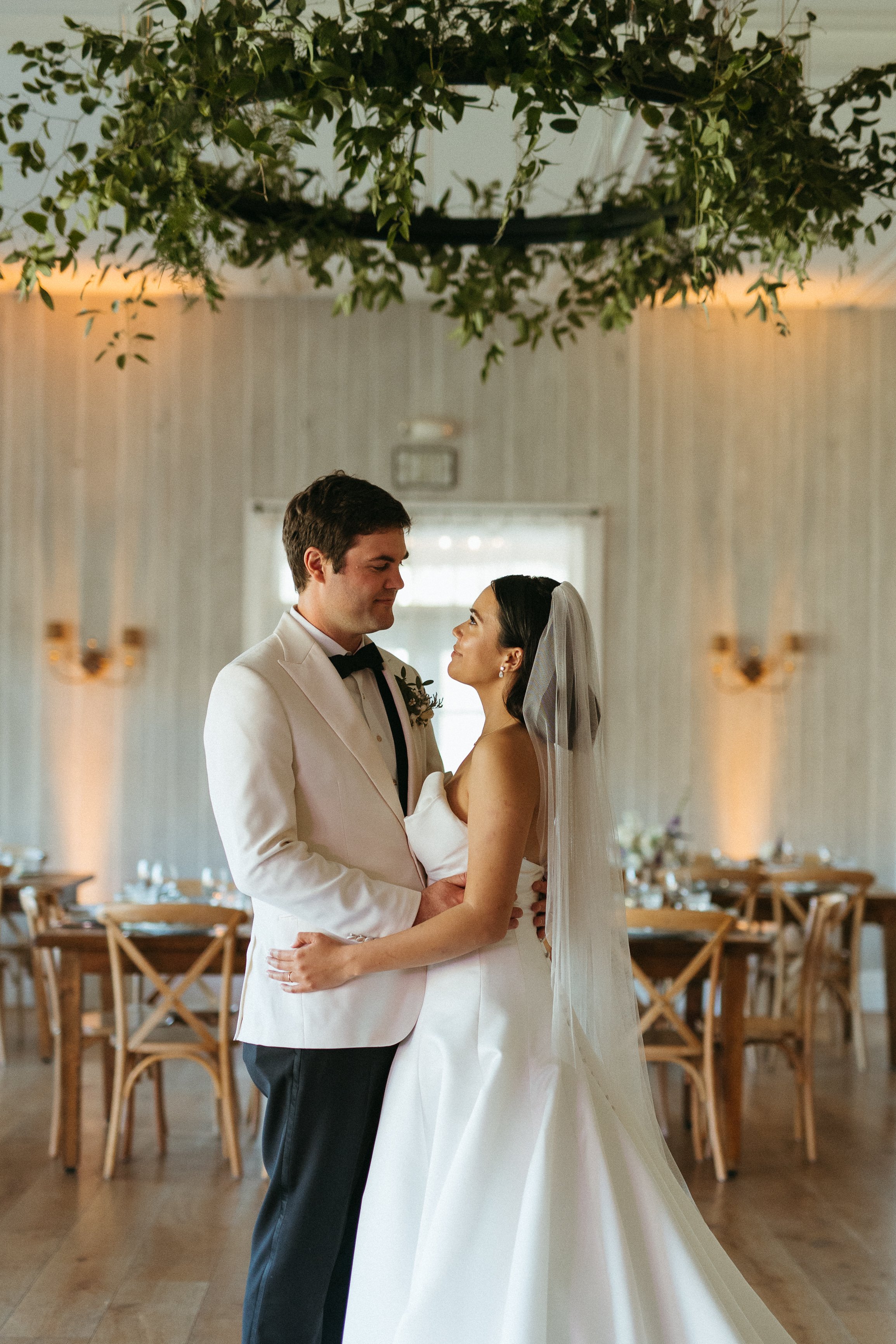 A bride and groom standing close together indoors during their wedding ceremony, gazing into each other's eyes, with decorated tables and chairs in the background.