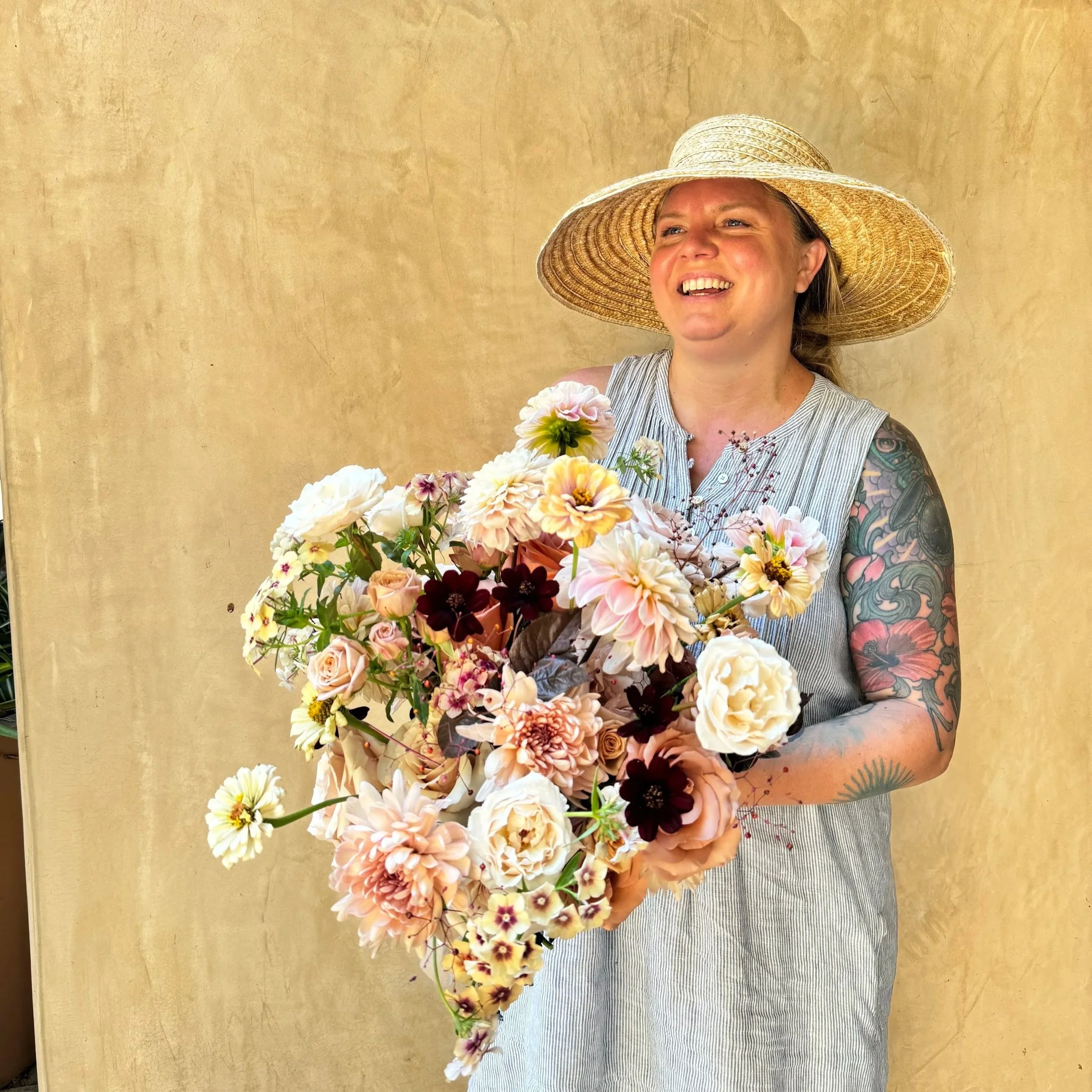 A woman wearing a large straw hat and a sleeveless striped top holding a large bouquet of mixed flowers, smiling, with a yellow wall in the background.