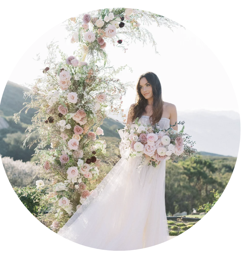 A woman in a white wedding dress holding a bouquet of pink and white flowers, standing outdoors beside a large floral arrangement arch made of pink, white, and purple blooms, with a scenic background of green hills and a mountain in the distance.