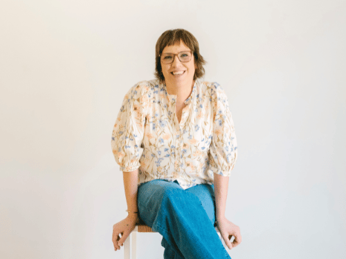 A woman with glasses and short hair, wearing a floral blouse and blue jeans, smiling while sitting on a white stool against a plain white background.