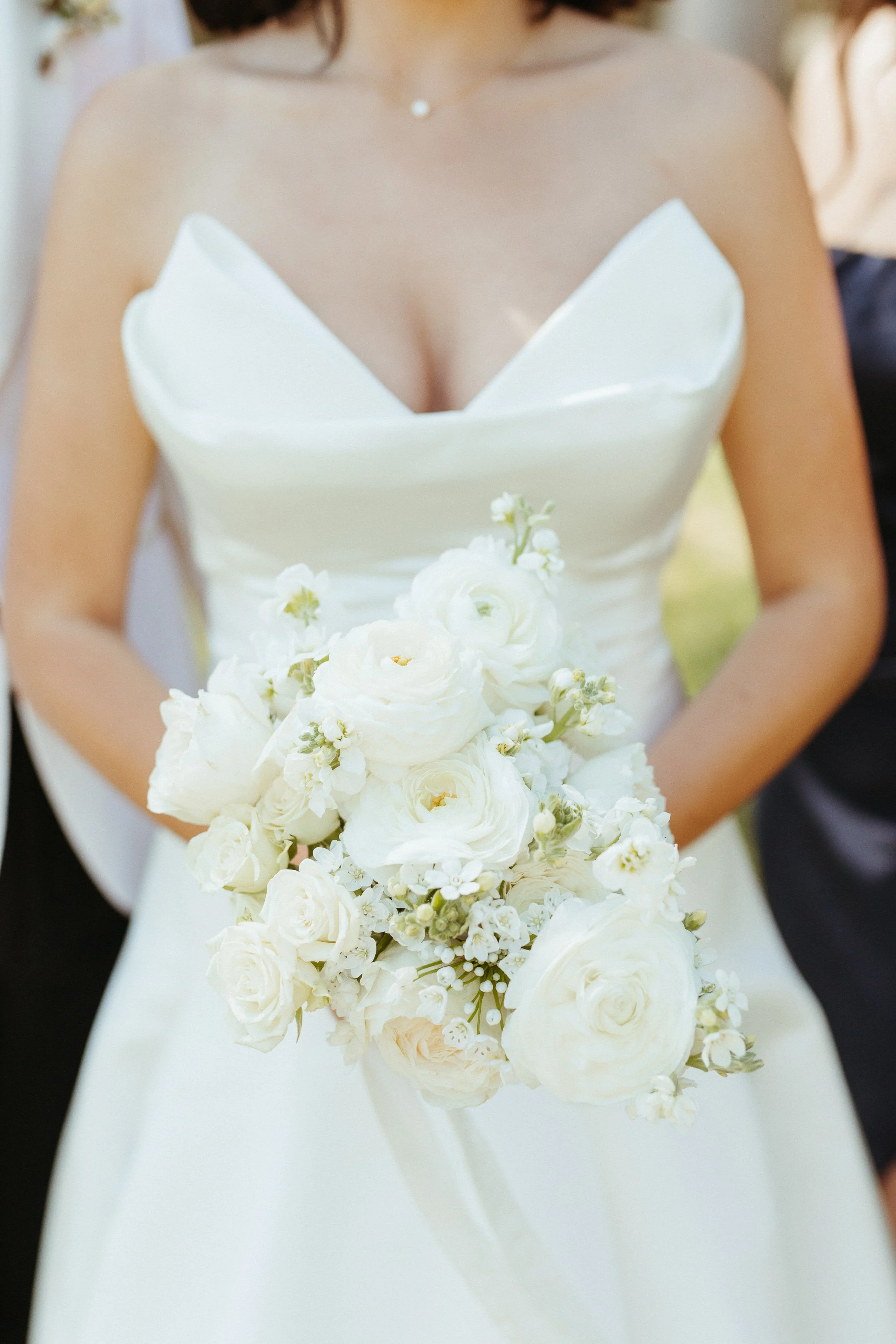 A bride in a strapless white wedding gown holding a large bouquet of white flowers, including roses and ranunculus, during a wedding ceremony.