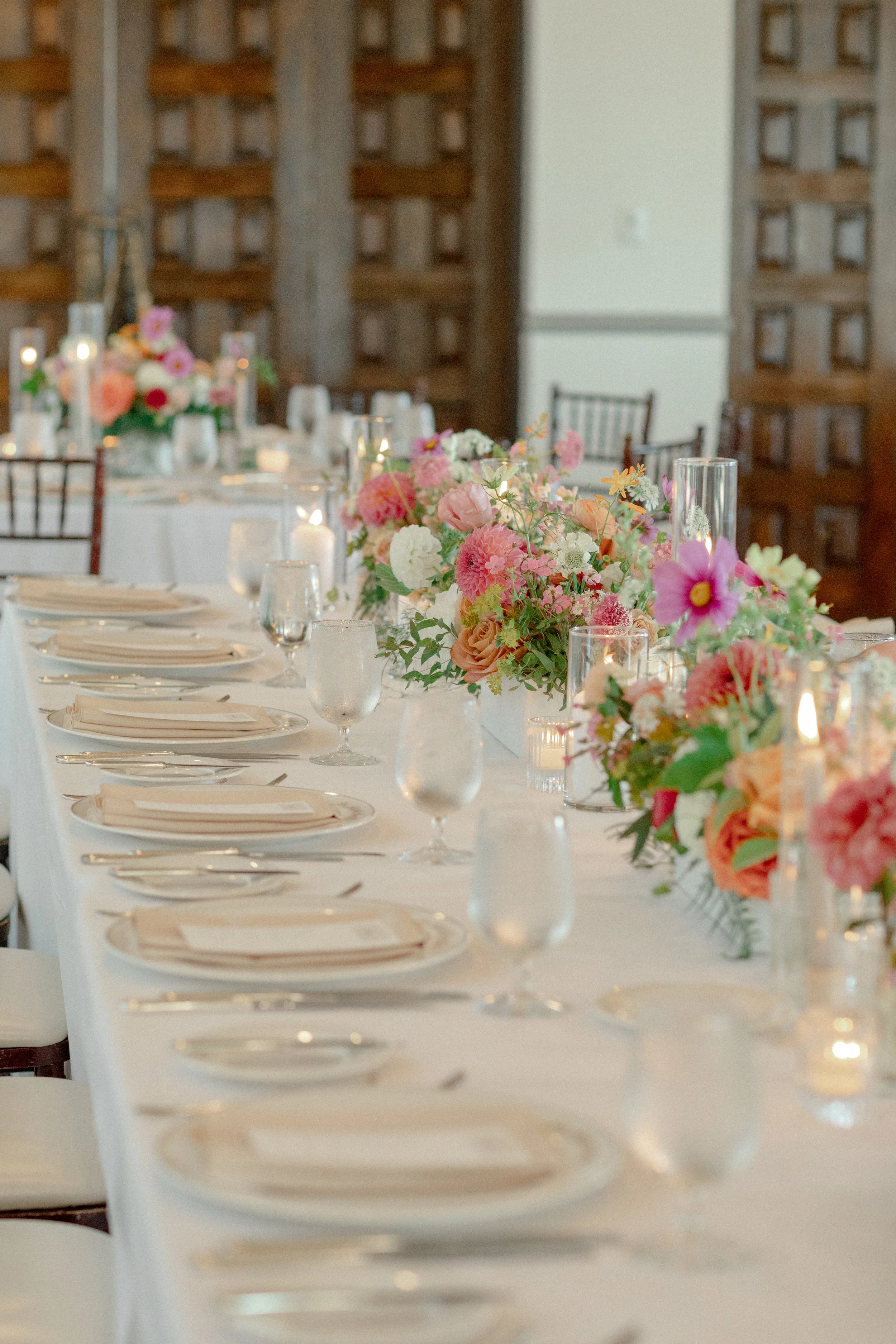 A long banquet table decorated with pink, white, and peach flowers in vases, with glass candle holders, wine glasses, and plates set for a formal event in a room with wooden lattice walls.
